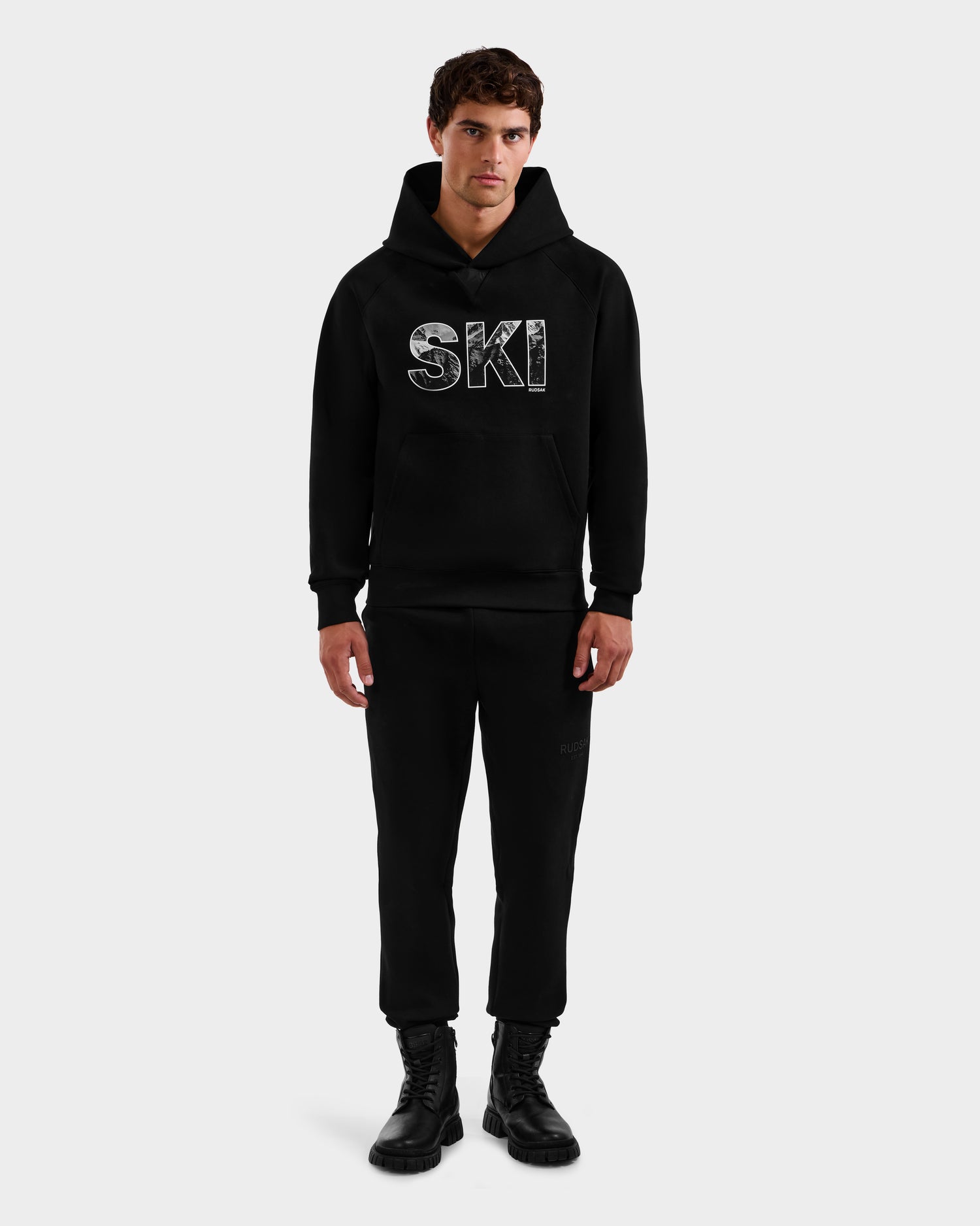 A man faces forward against a white background, wearing the Rudsak MIKAEL - 6525647 Black men’s ski hoodie with SKI in large white letters, paired with black pants and boots.