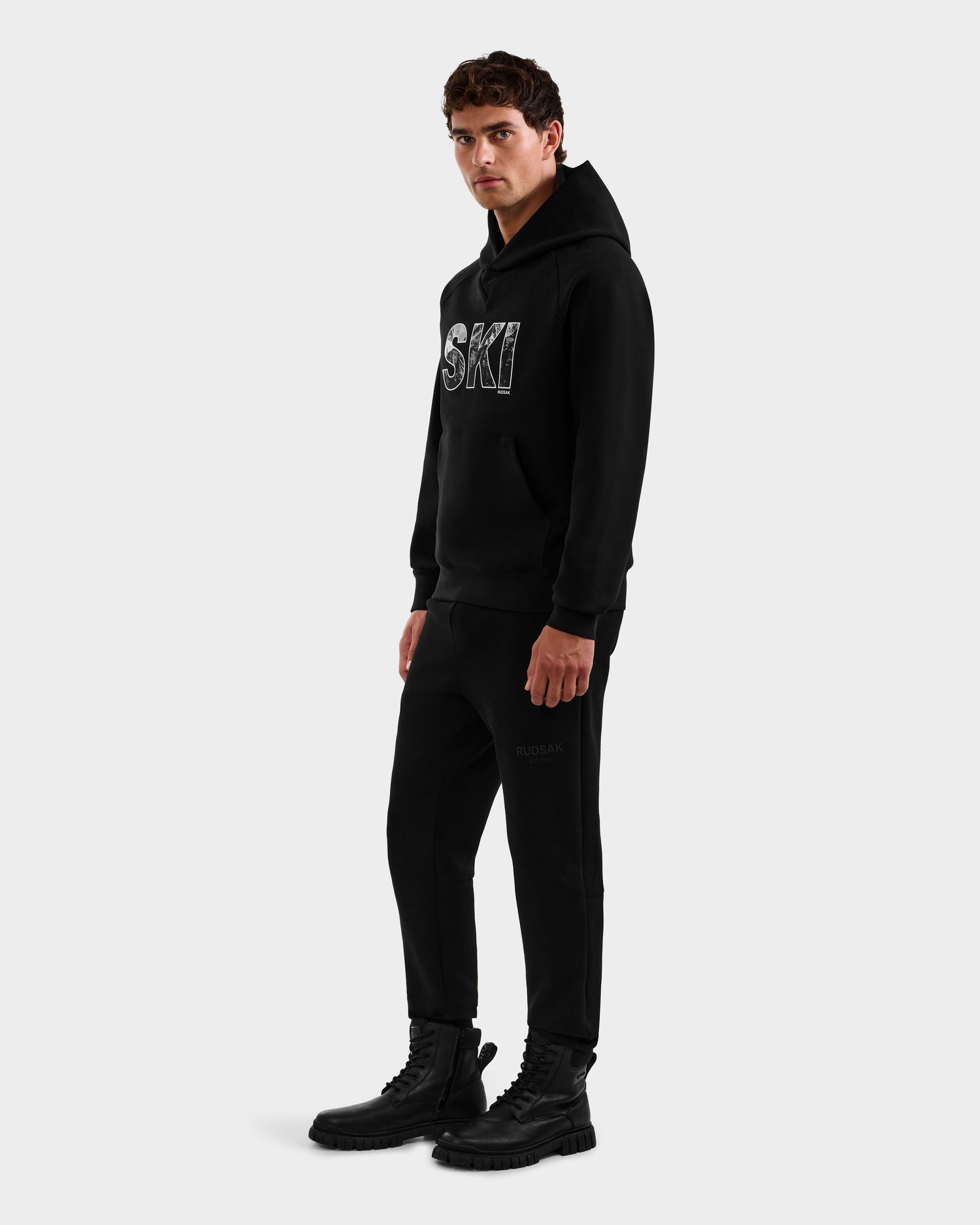 A man stands against a plain light gray background wearing the Rudsak MIKAEL - 6525647 Black men’s ski hoodie with "SKI" in white, paired with black jogger pants and black lace-up boots.