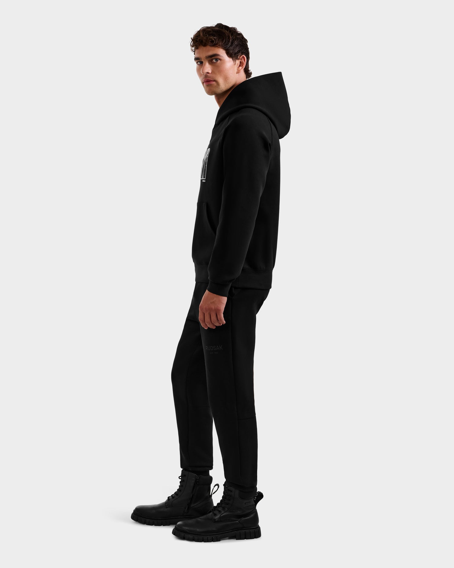 A man models the Rudsak MIKAEL - 6525647 Black men’s ski hoodie, black jogger pants, and black boots, standing sideways and glancing at the camera against a plain white background.