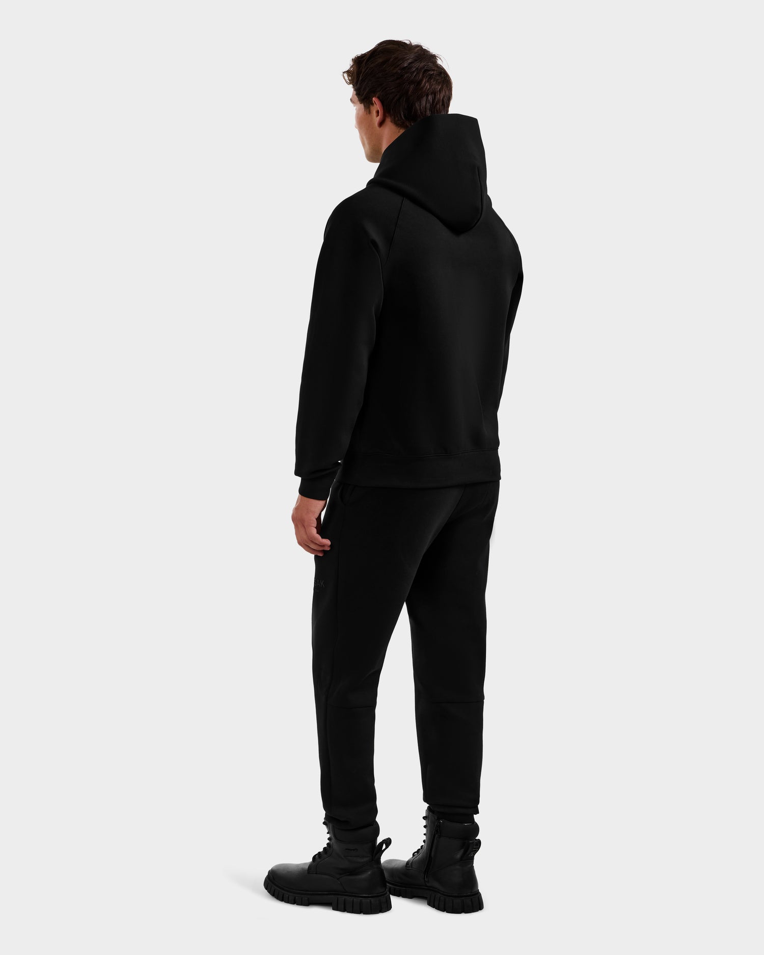 A man faces away from the camera, wearing the Rudsak MIKAEL - 6525647 Black men’s ski hoodie with black pants and boots, set against a plain light gray background.
