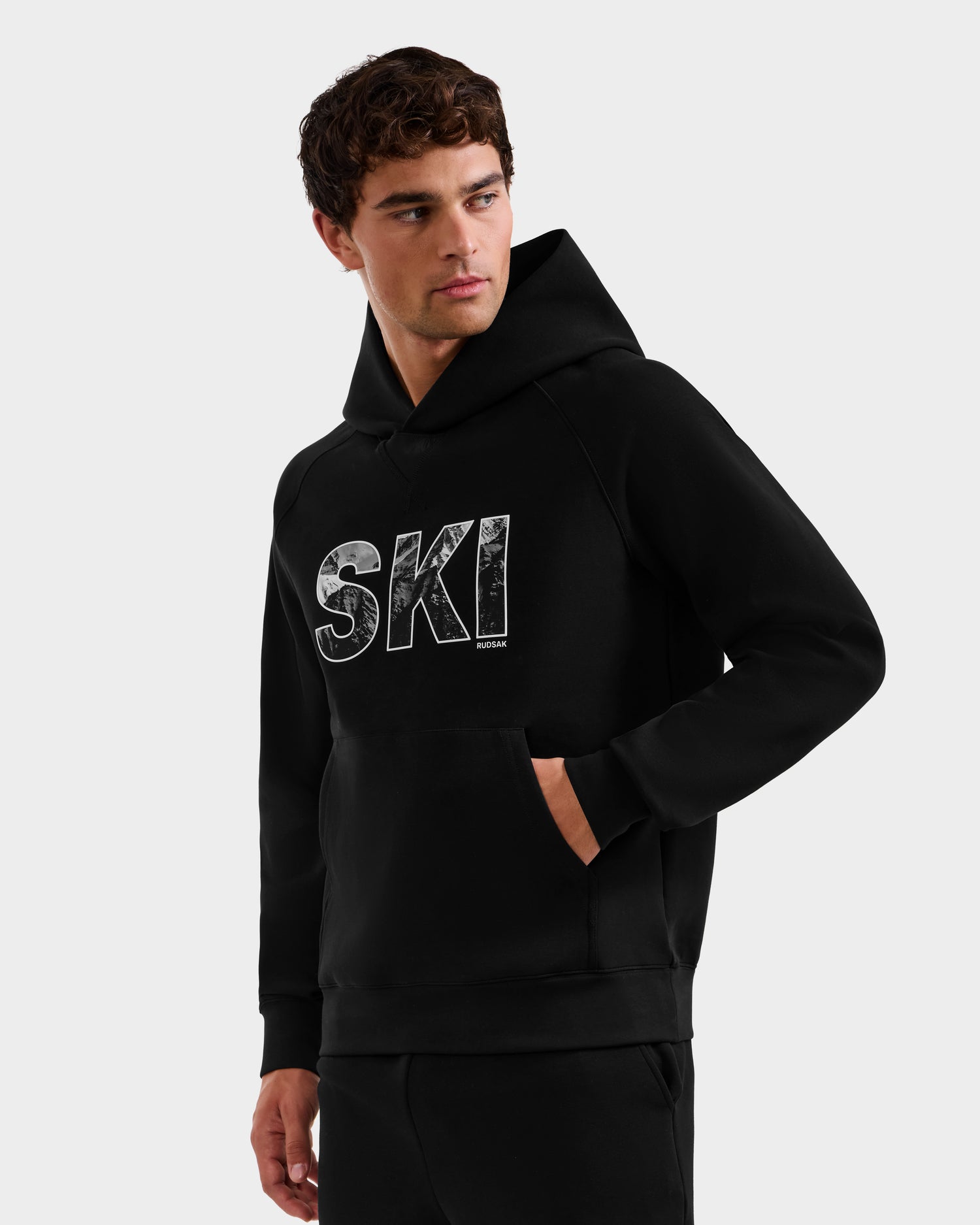 A man wearing the Rudsak MIKAEL - 6525647 Black men’s ski hoodie with large, distressed white “SKI” lettering on the front stands with one hand in his pocket, looking to the side.