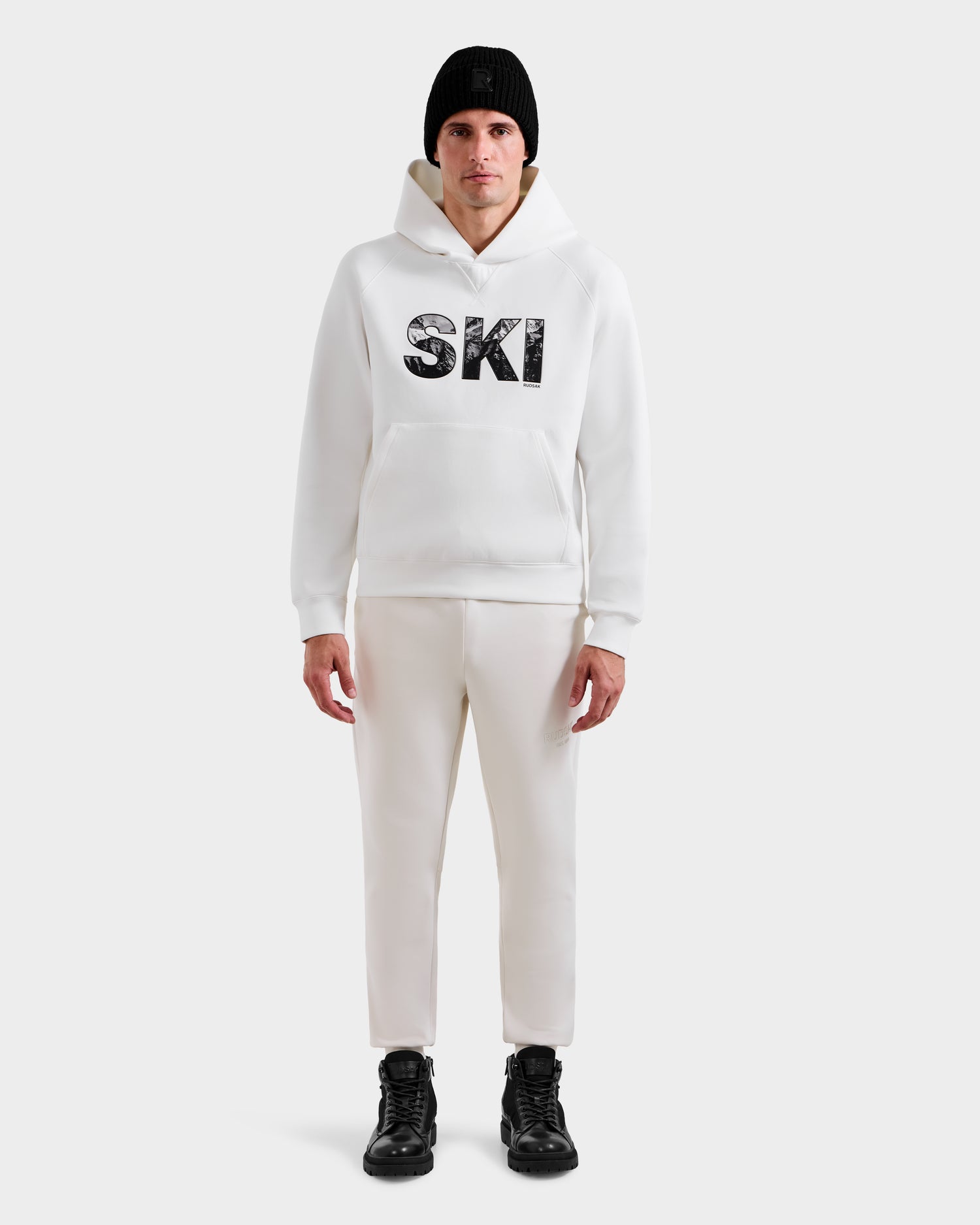 A person faces forward against a plain light background, wearing the Rudsak MIKAEL - 6525647 Iceland men’s ski hoodie, a black beanie, white pants, and black boots.