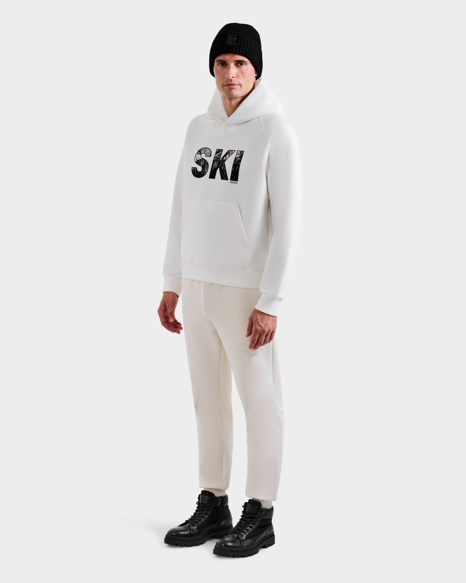 A person wears the Rudsak MIKAEL - 6525647 Iceland men’s ski hoodie in white, paired with matching white pants, black lace-up boots, and a black knit beanie, standing against a plain white background.
