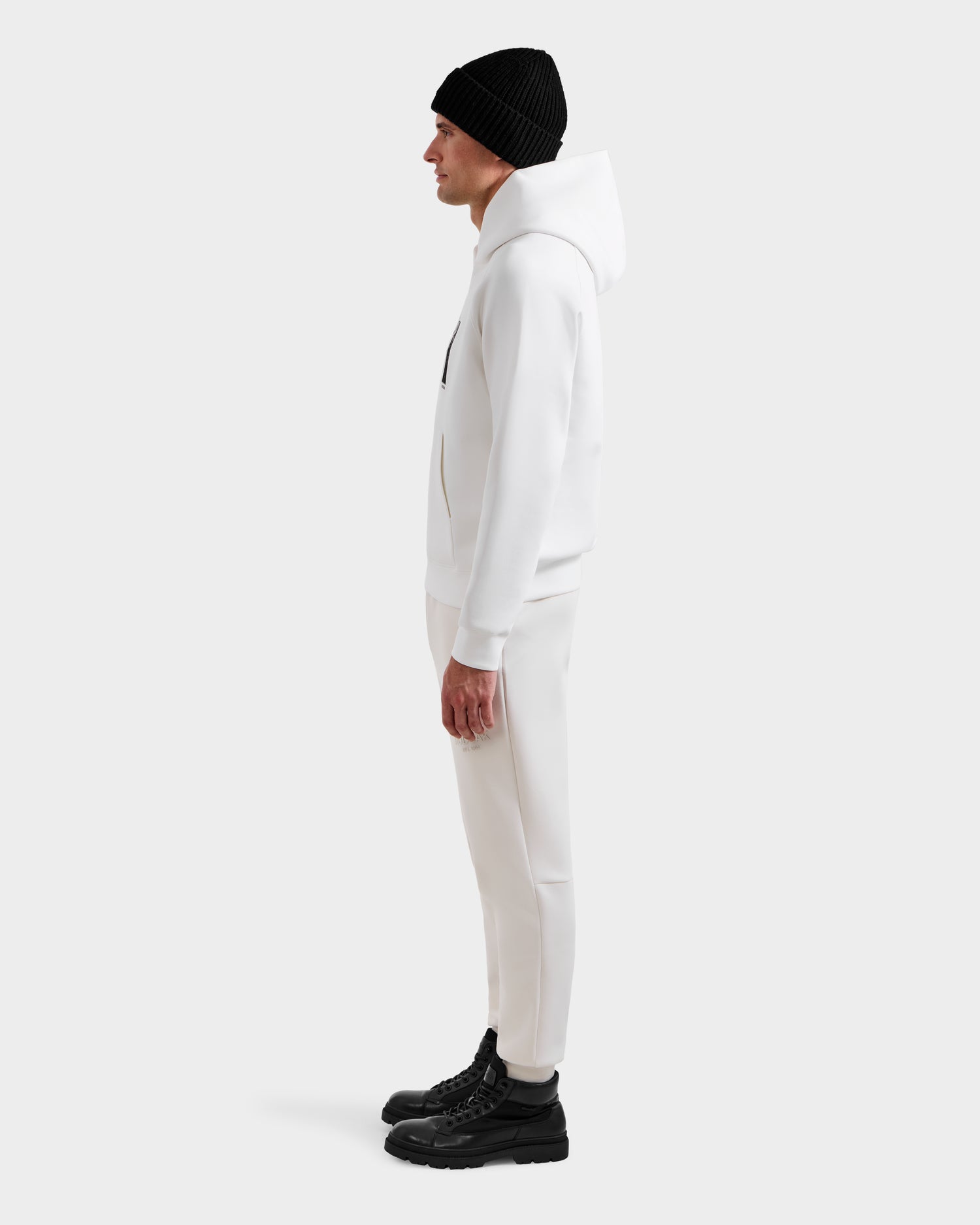 A man stands in profile against a plain background, wearing the Rudsak MIKAEL - 6525647 Iceland ski hoodie in white, with matching pants, a black beanie, and black boots.