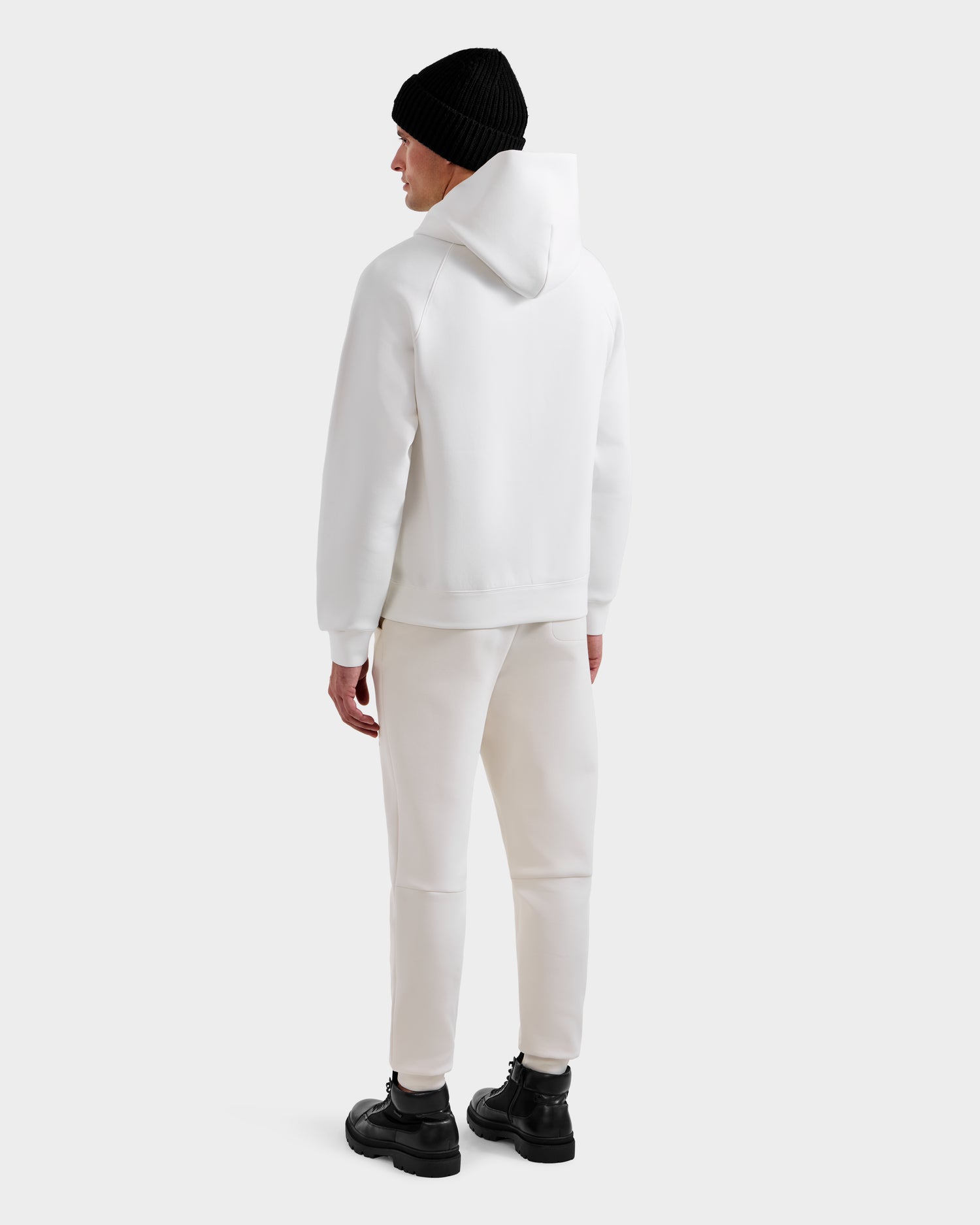 A person faces away wearing Rudsak's MIKAEL - 6525647 Iceland men’s ski hoodie, paired with matching white sweatpants, a black beanie, and black boots against a plain light background.