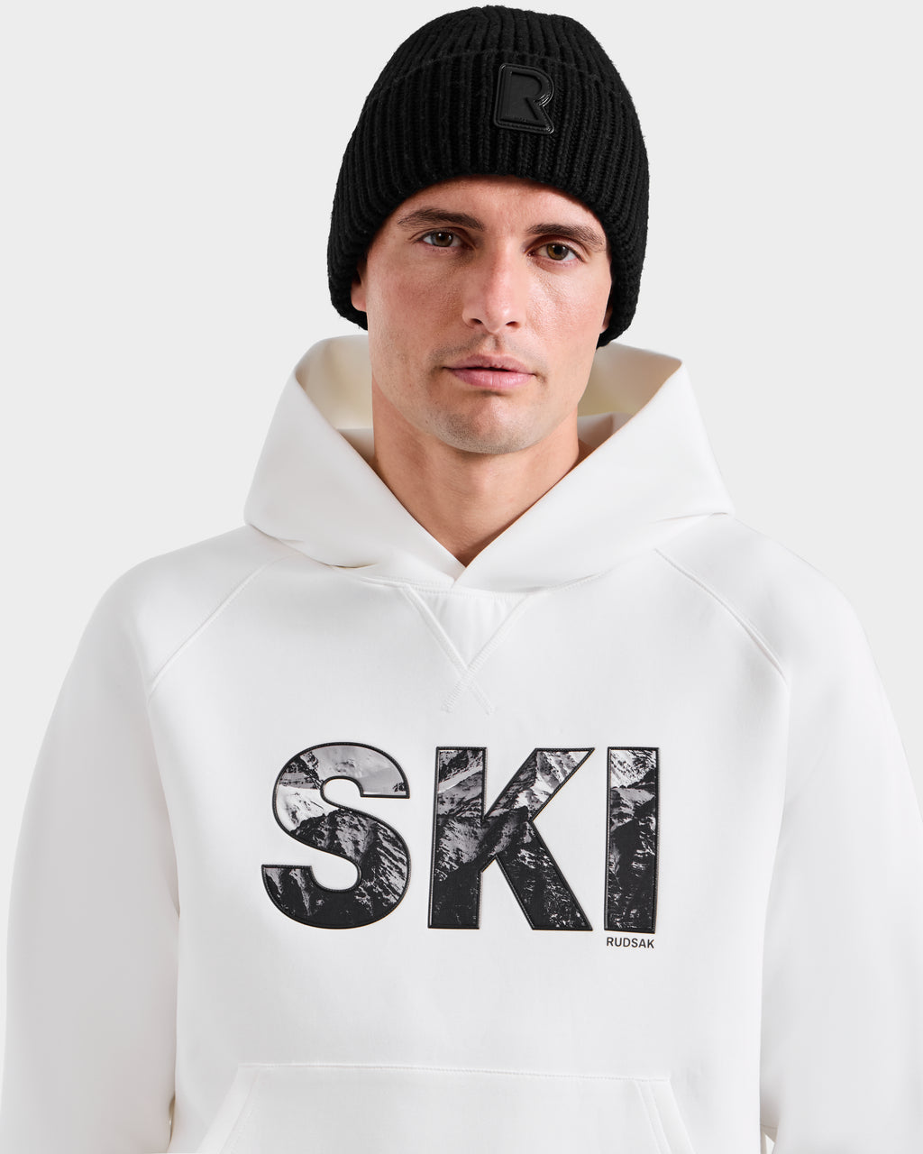 A man wearing a black knit beanie and the Rudsak MIKAEL - 6525647 Iceland premium cotton men’s ski hoodie with bold “SKI” text stands against a plain light background, looking directly at the camera.