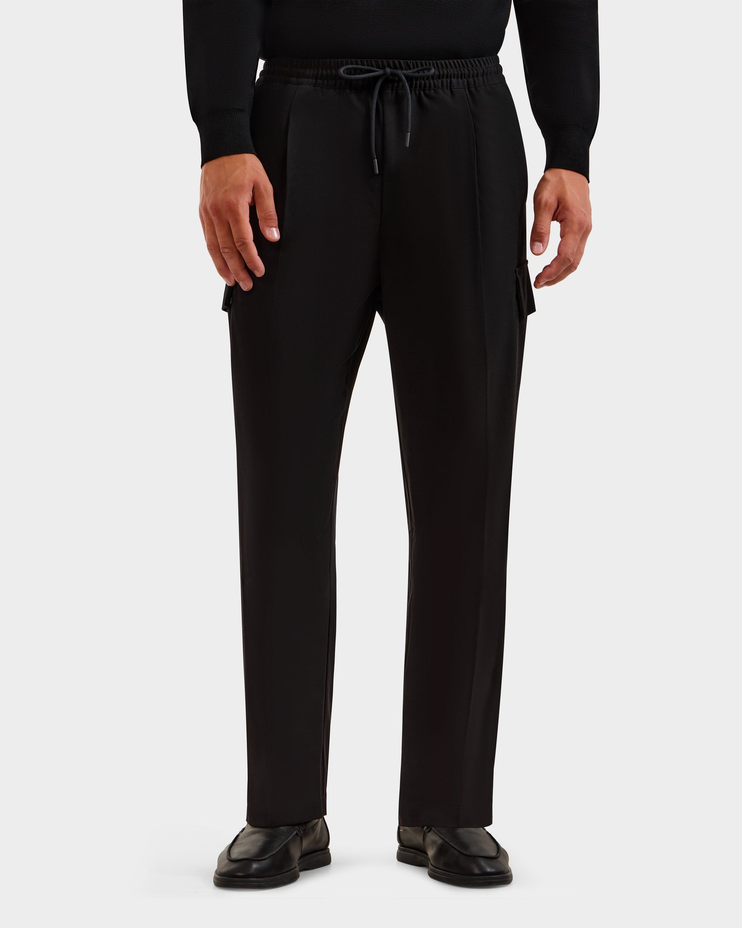 A person wears Rudsak’s MIKEY - 6525597 Black men’s trousers with a relaxed fit and side pockets, styled with a black long-sleeve shirt and shoes, standing against a plain white background.