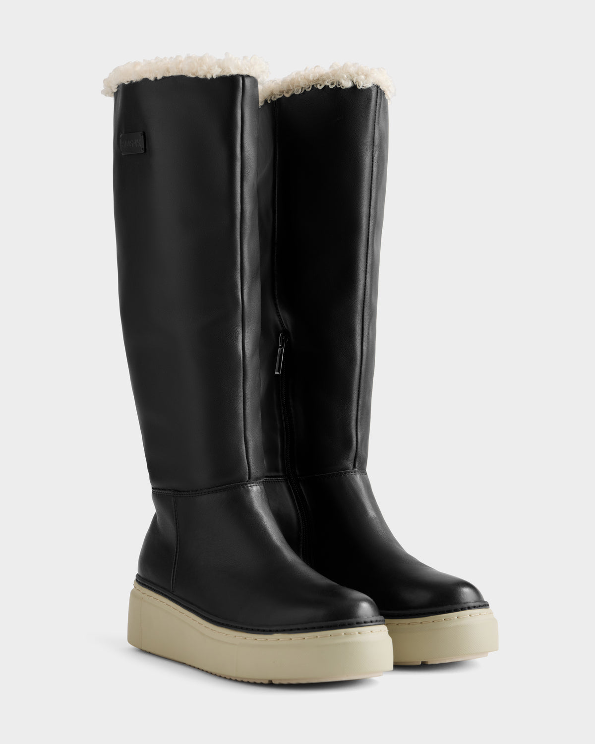The Rudsak MIKIE L - 8225580 Black features women's black leather knee-high boots with white platform soles and visible cream faux fur lining at the top, displayed against a plain light gray background.