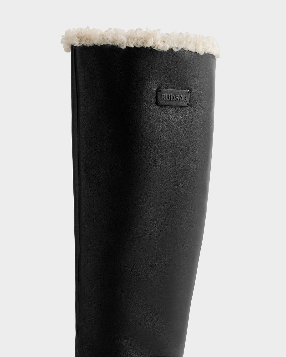 A close-up of the Rudsak MIKIE L - 8225580 Black winter boot, showcasing its leather exterior, shearling-lined top, and signature brand logo patch.