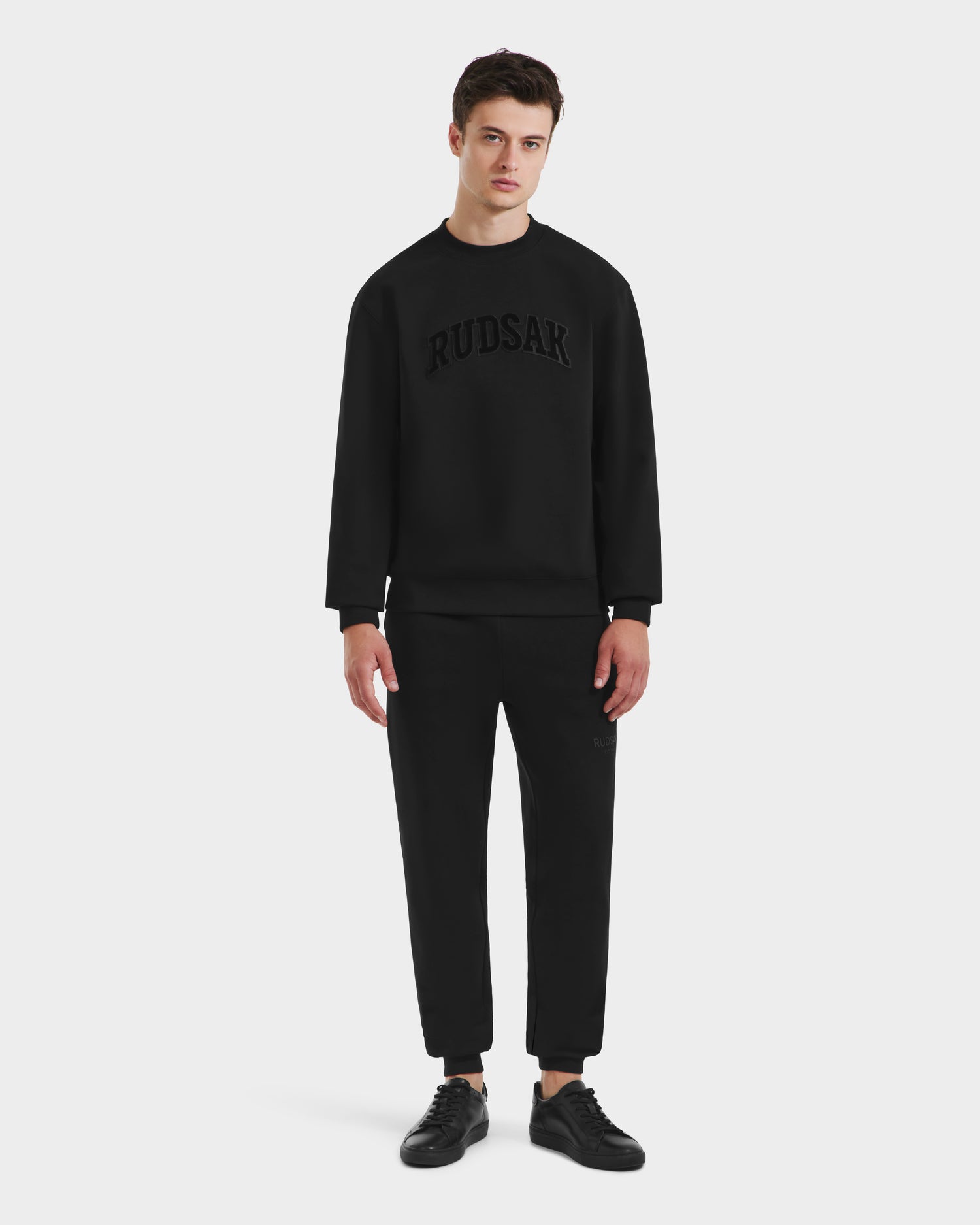 A young man faces forward against a white background, wearing the Rudsak MILOS - 6525646 Black men’s premium cotton blend crewneck sweatshirt with matching black pants and sneakers.