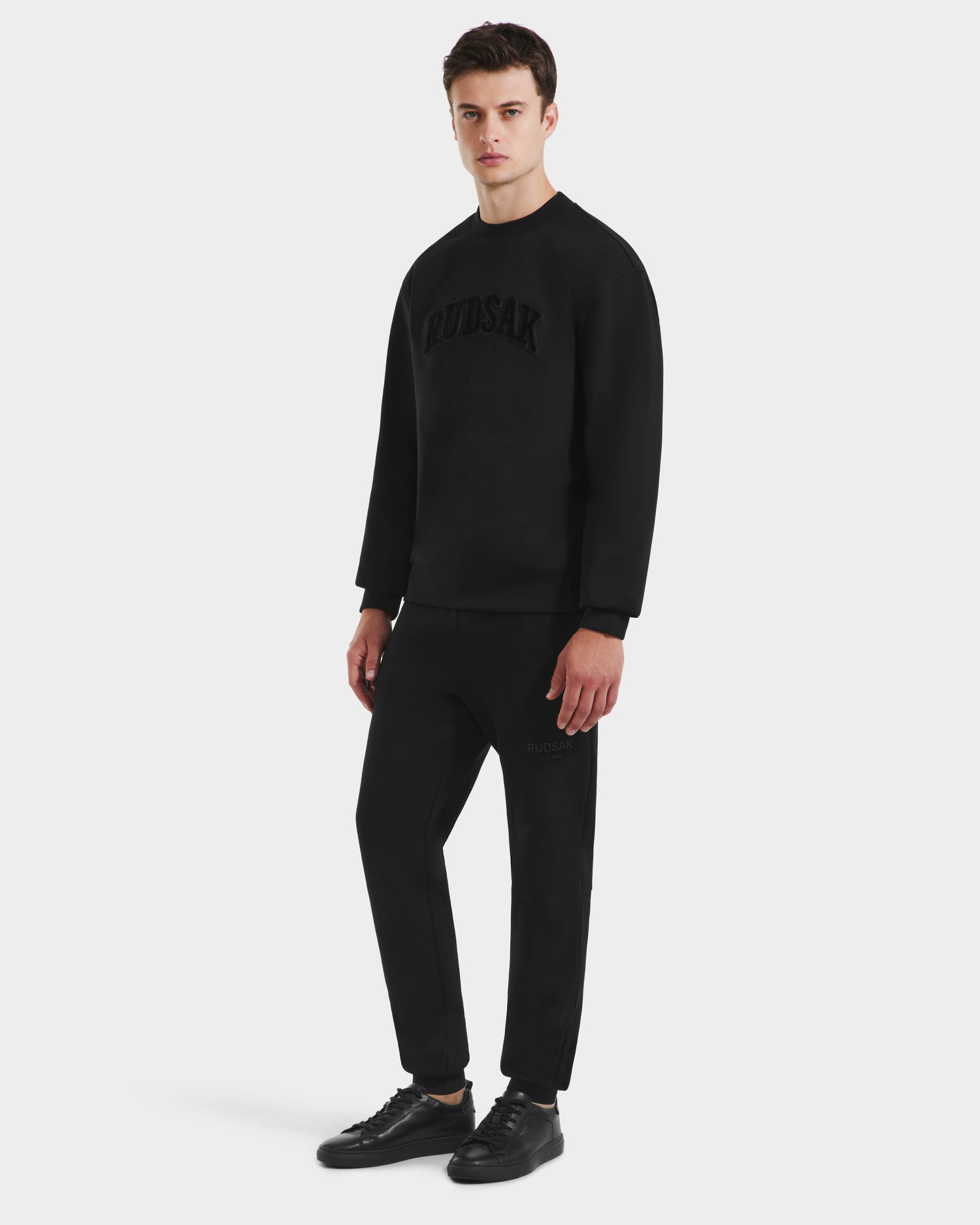 A man stands against a white background, wearing the Rudsak MILOS – 6525646 Black men’s premium cotton blend crewneck sweatshirt with matching black jogger pants and black sneakers.