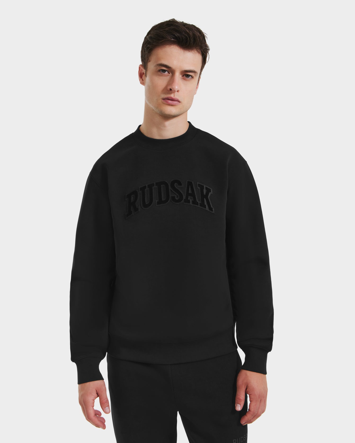 A young man stands against a white background, wearing Rudsak’s MILOS - 6525646 Black sweatshirt and black pants. The premium crewneck features “RUDSAK” in bold, embossed letters across the chest.