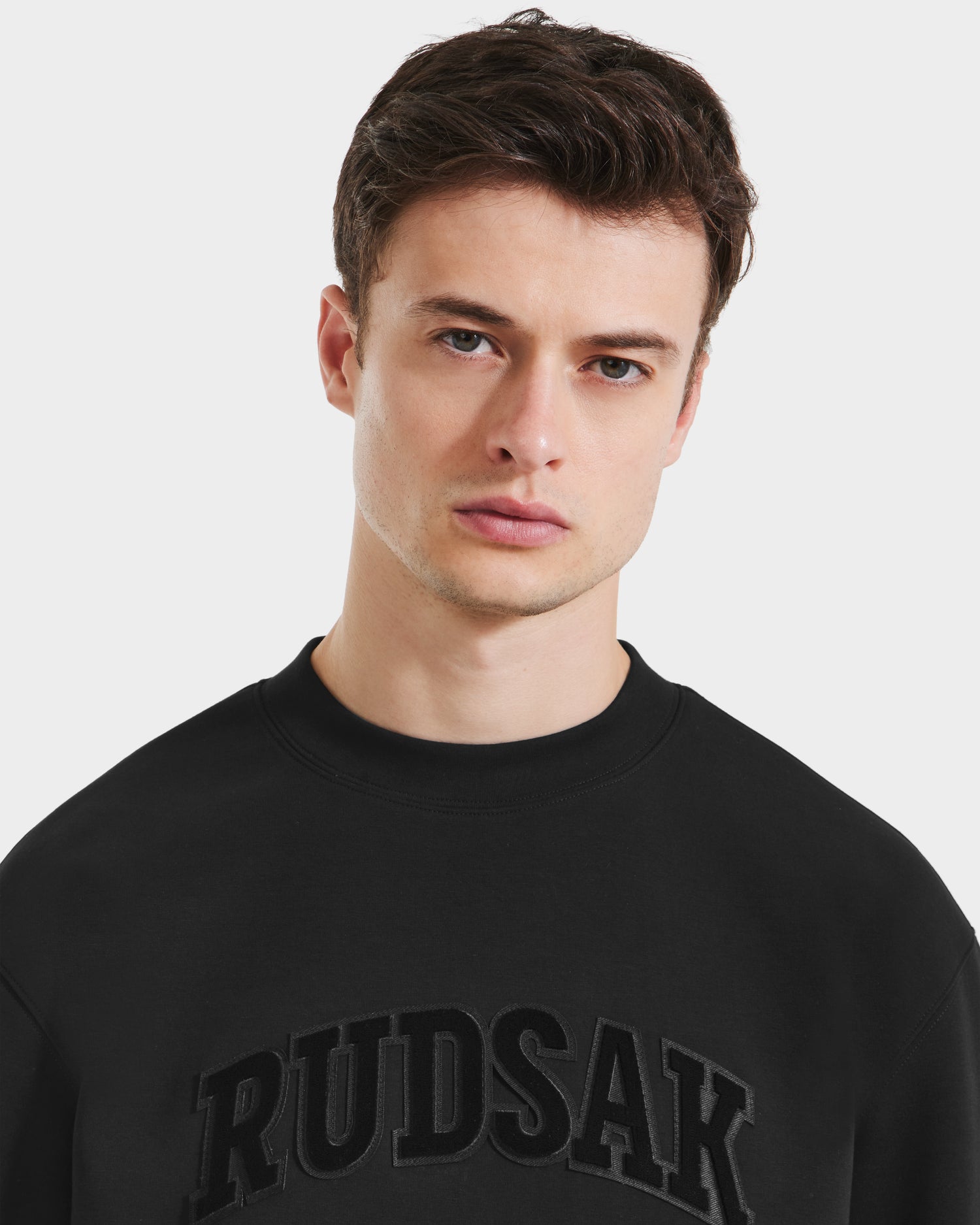 A young man with short brown hair wears the Rudsak MILOS - 6525646 Black men’s crewneck sweatshirt with bold RUDSAK lettering on the chest, looking directly at the camera with a neutral expression.