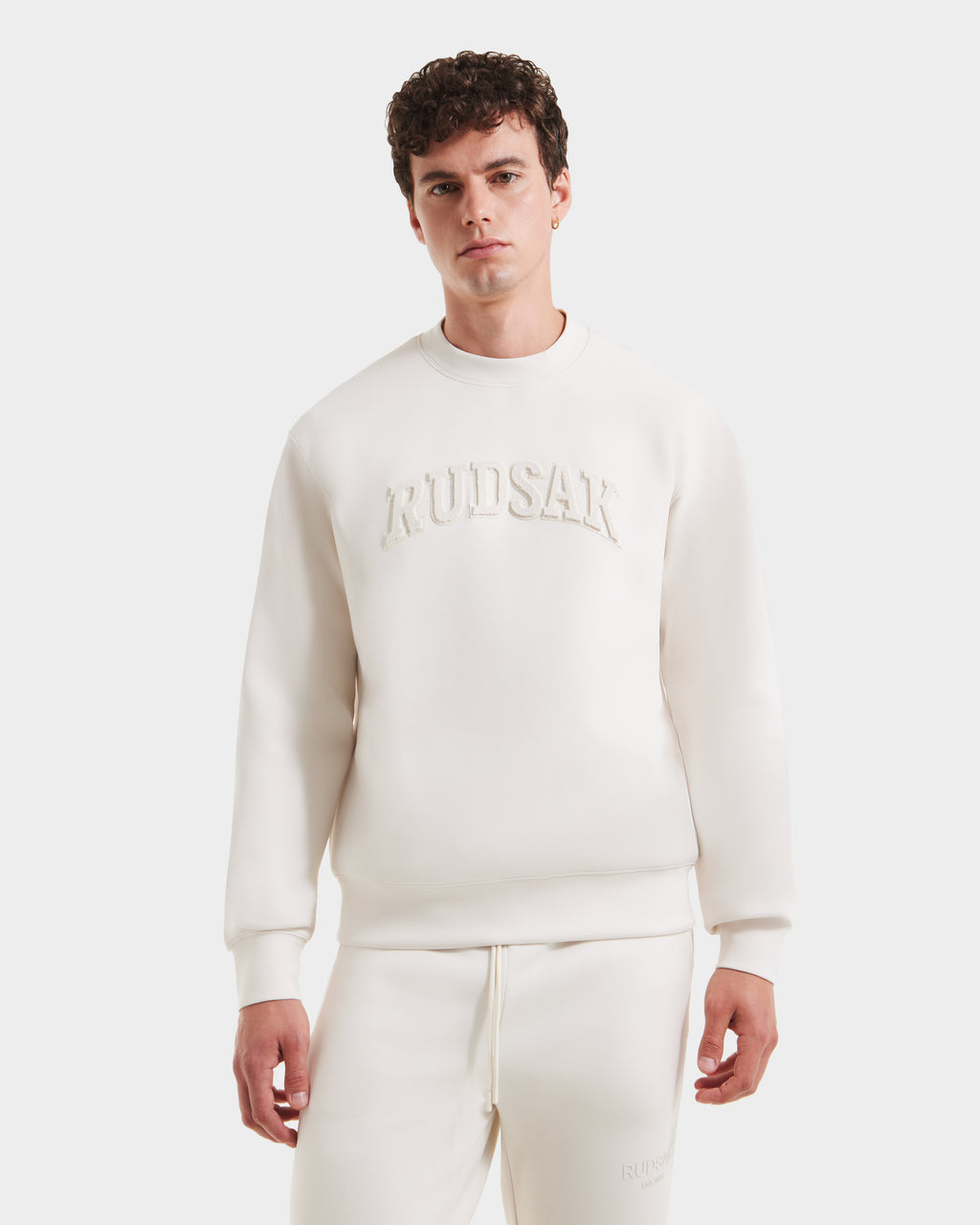 A young man in the MILOS - 6525646 Vanilla men’s premium Rudsak sweatshirt and matching sweatpants set with embossed Rudsak text stands against a plain white background.