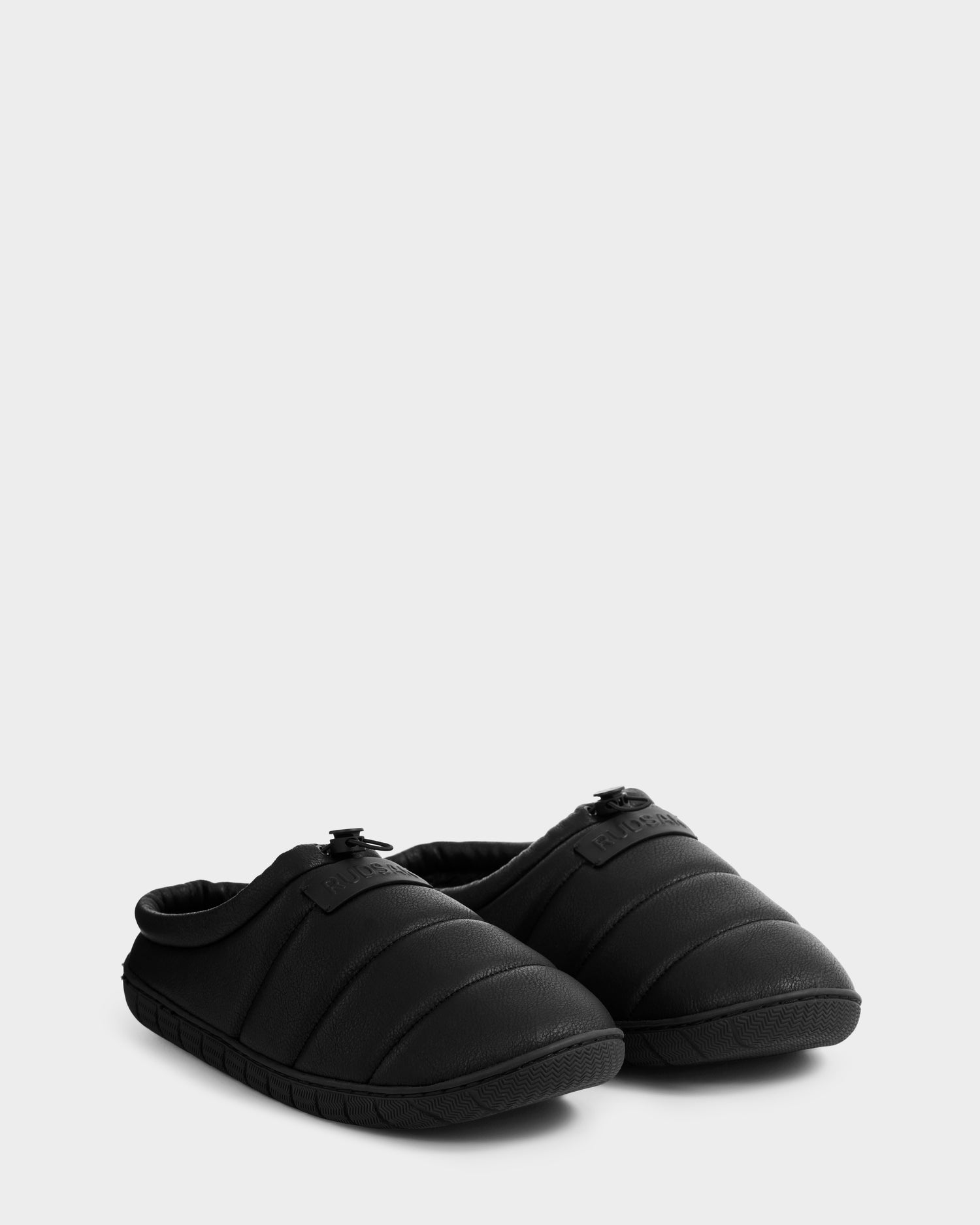 A pair of Rudsak MILTON - M225504 Black men’s quilted slippers with rounded toes, cozy faux-shearling lining, and textured rubber soles is displayed side by side on a light gray background.