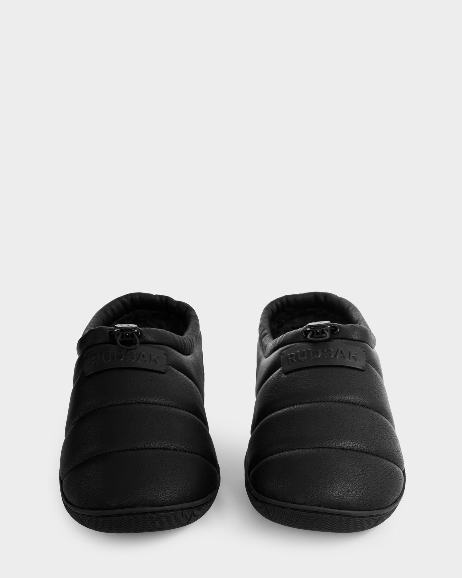 The Rudsak MILTON - M225504 Black men's quilted slippers, featuring visible stitching, a rounded toe, and cozy faux-shearling lining, are pictured side by side on a plain light background.