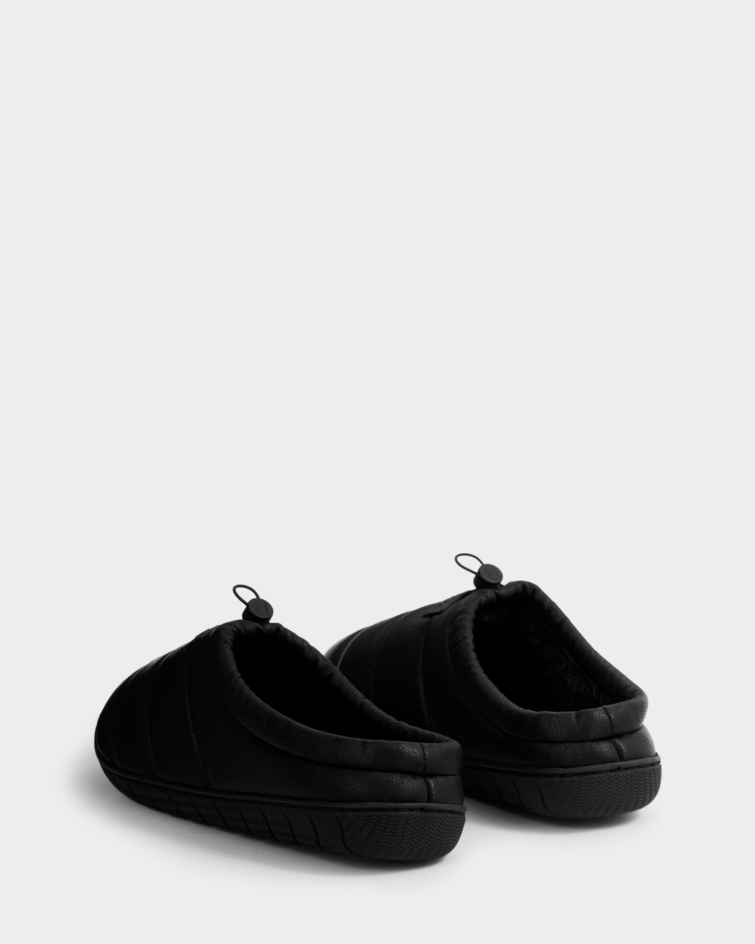 The Rudsak MILTON - M225504 Black men’s quilted slip-on slippers in vegan leather feature pull tabs at the back, shown from an angle highlighting the heels and sides against a plain light gray background.