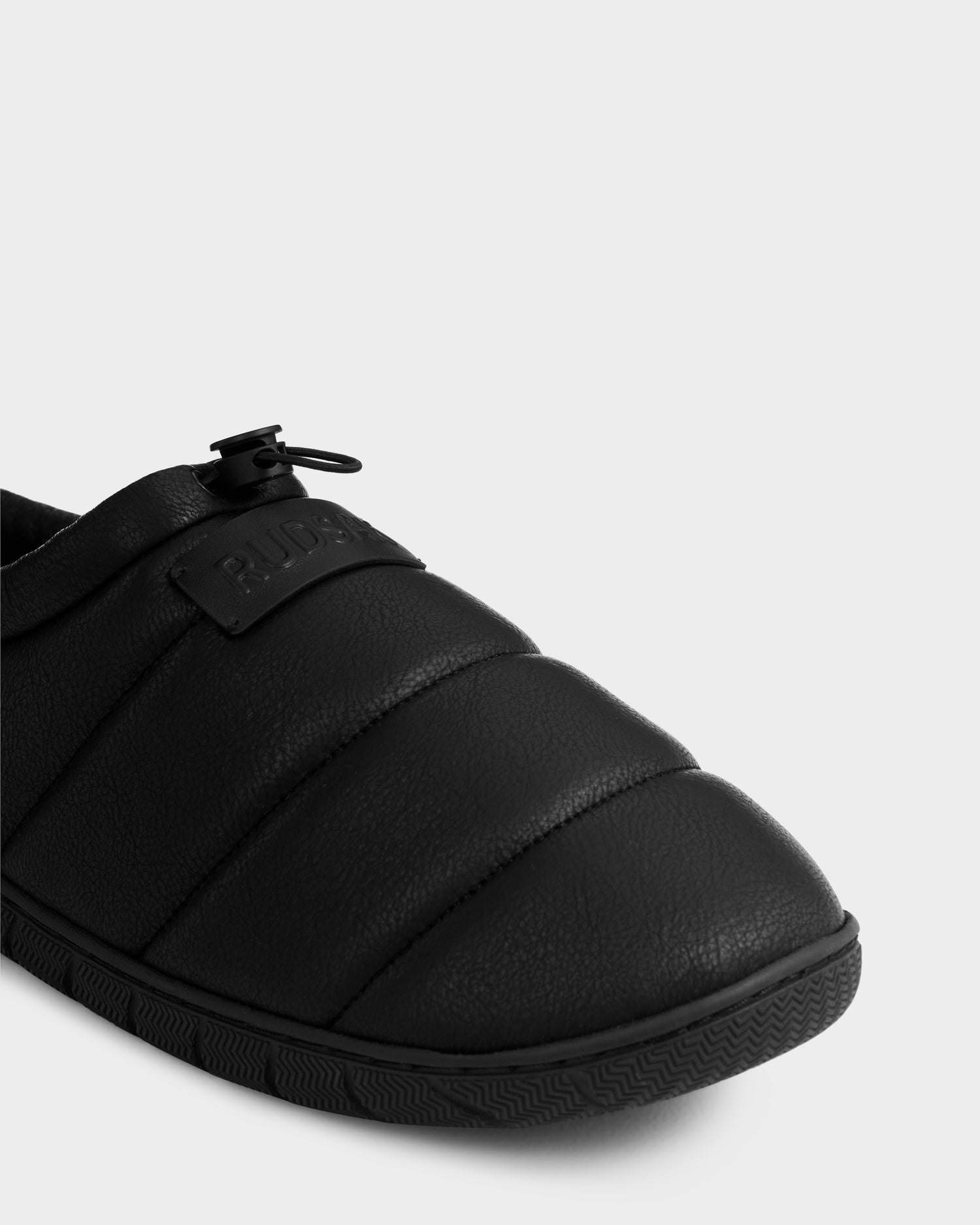 Close-up of the Rudsak MILTON - M225504 Black men's quilted slipper, featuring a padded upper, textured rubber sole, drawstring toggle, faux-shearling lining for comfort, and an embossed brand label on the upper.