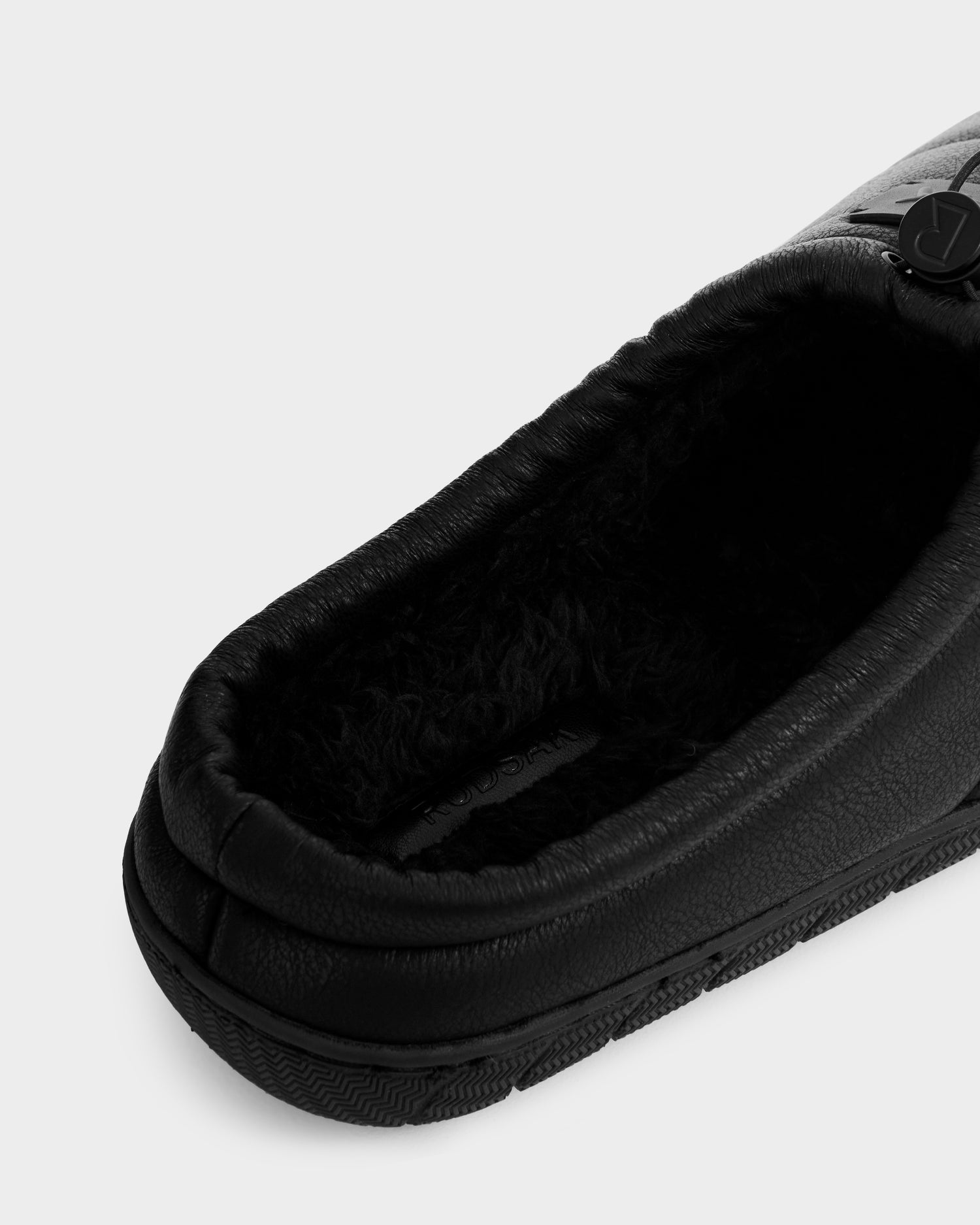 Close-up of the Rudsak MILTON - M225504 Black slipper, featuring black vegan leather, a textured sole, and soft faux-shearling lining, shown overhead against a plain white background.