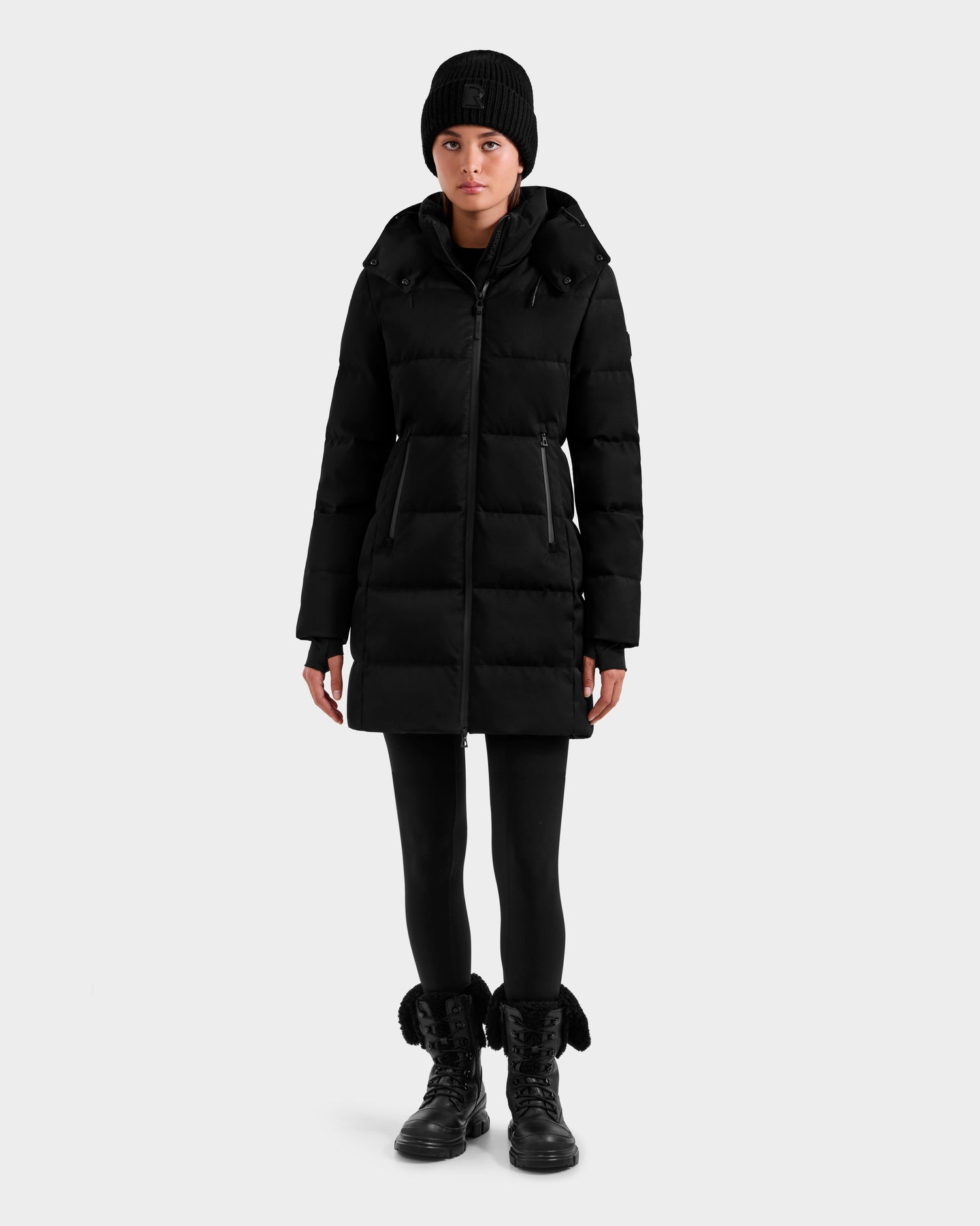 A person wears the Rudsak MIRIAM MW - L125604 Black long women's down puffer coat with RDS-certified down, styled with a black beanie, black leggings, and lace-up boots, facing forward against a plain light gray background.