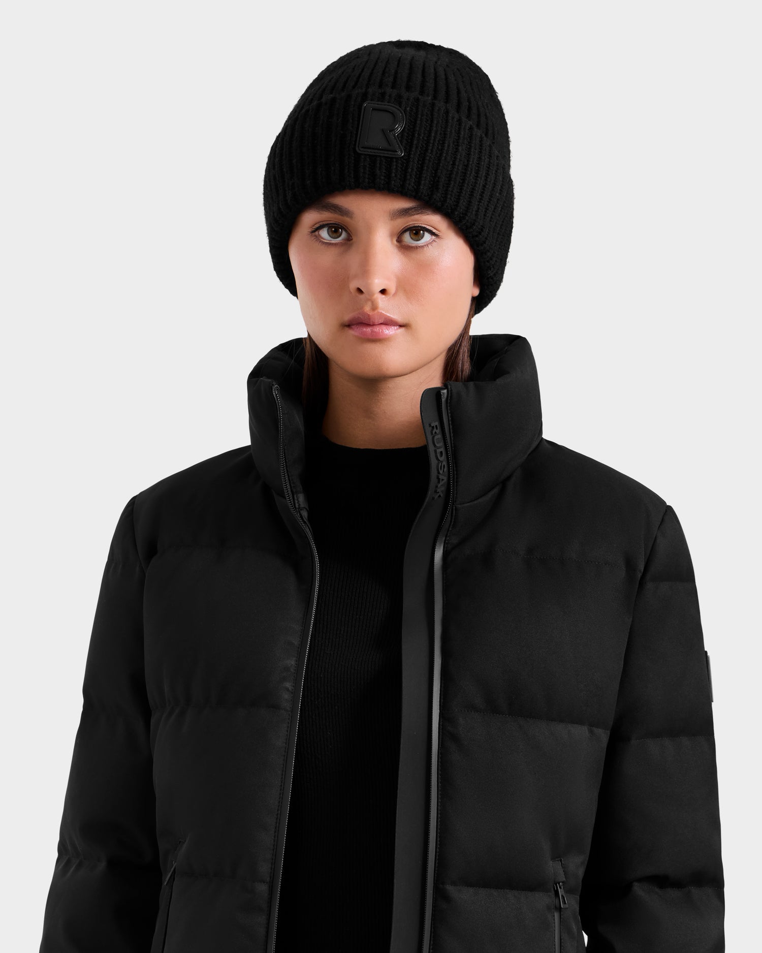 A person wearing the Rudsak MIRIAM MW - L125604 Black RDS-certified down puffer jacket with a high collar and a knit beanie stands against a light gray background, creating an essential winter look.