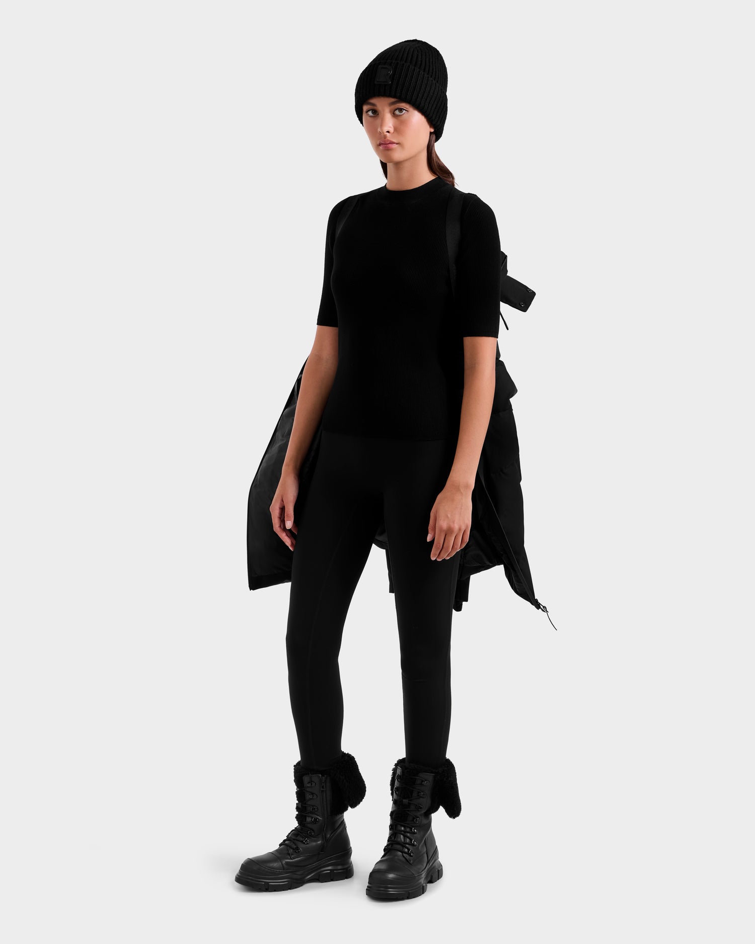 A woman wears a black beanie, short-sleeve top, leggings, boots, and the Rudsak MIRIAM MW - L125604 Black RDS-certified down jacket draped over her shoulders against a plain light gray background—perfect for winter.