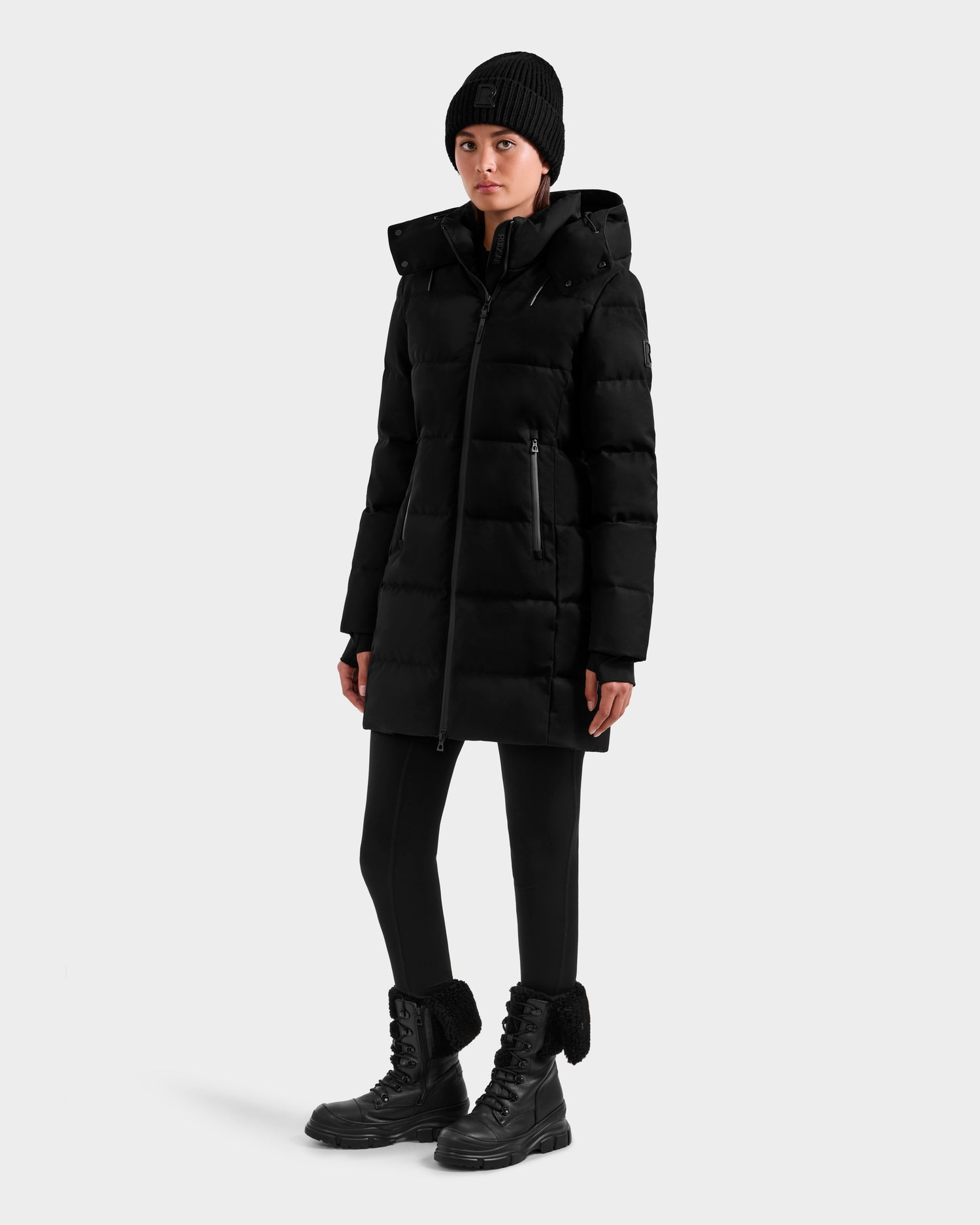 A person wearing the Rudsak MIRIAM MW - L125604 Black down puffer coat, paired with a black knit beanie, black leggings, and black lace-up winter boots stands slightly turned to the side against a plain light background.