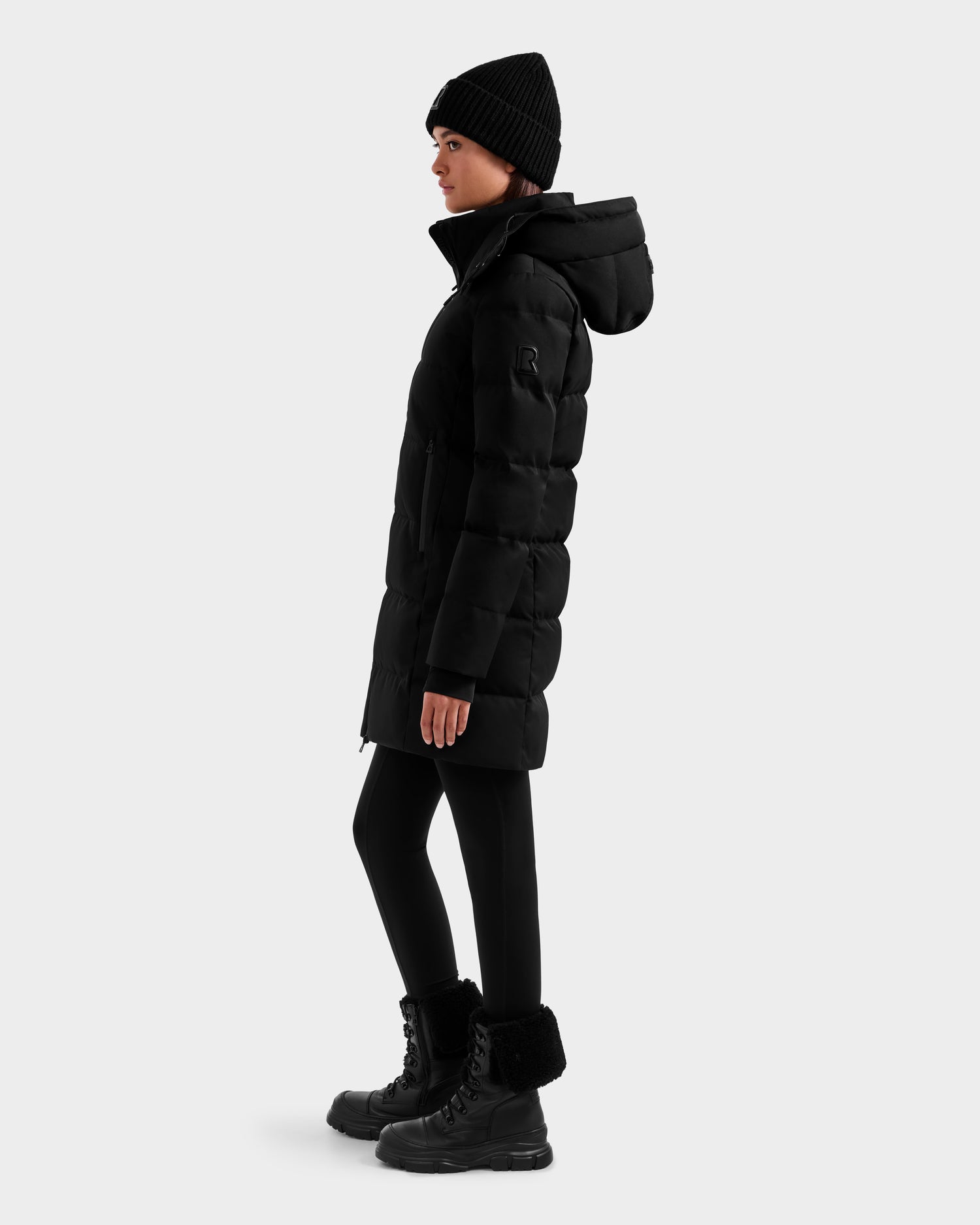 A person stands sideways against a plain light background in the Rudsak MIRIAM MW - L125604 Black women’s down puffer with RDS-certified down, paired with a black beanie, leggings, and winter boots—a chic winter wardrobe staple.