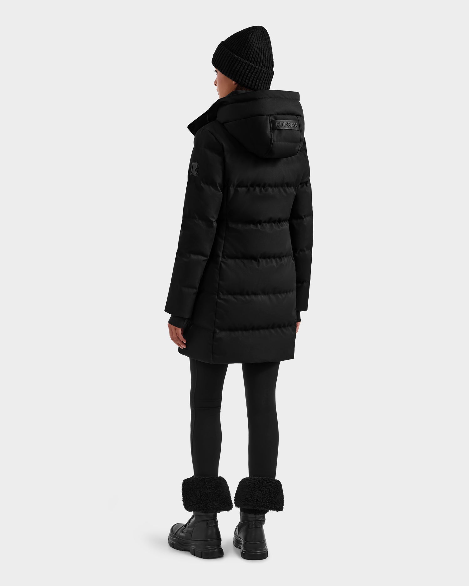 A person faces away, modeling the Rudsak MIRIAM MW - L125604 Black women’s down puffer with a hood, paired with black leggings, a beanie, and fur-cuffed boots against a plain light gray background.