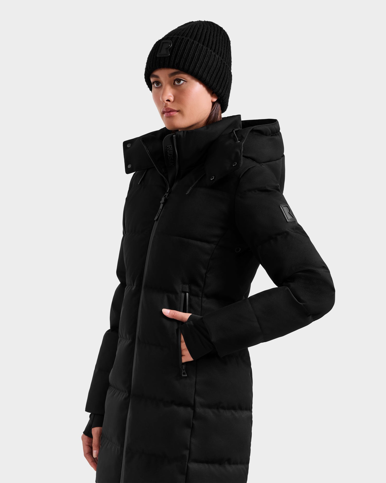 A woman wearing the Rudsak MIRIAM MW - L125604 Black down puffer coat with a hood and a black knit beanie stands with one hand in her pocket, looking to the side against a plain light background.