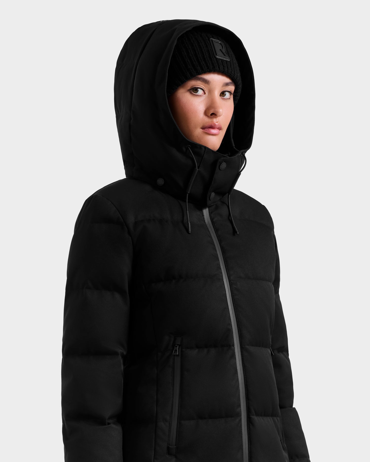 Wearing the Rudsak MIRIAM MW - L125604 Black down puffer and a black knit beanie, a person stands against a light background, looking to the side—an effortless addition to your winter wardrobe.