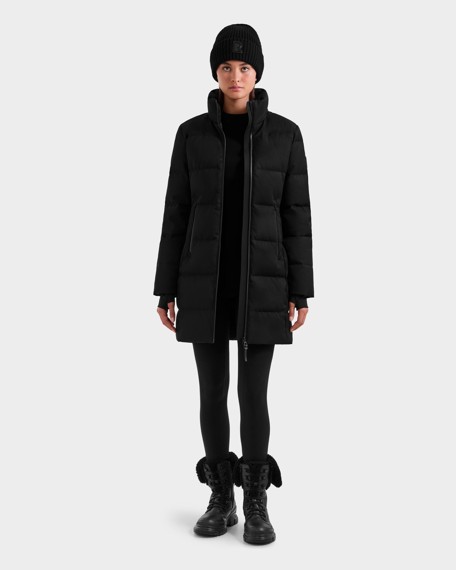 A person in the Rudsak MIRIAM MW - L125604 Black puffer coat, black beanie, leggings, and boots stands facing forward against a plain light gray background.