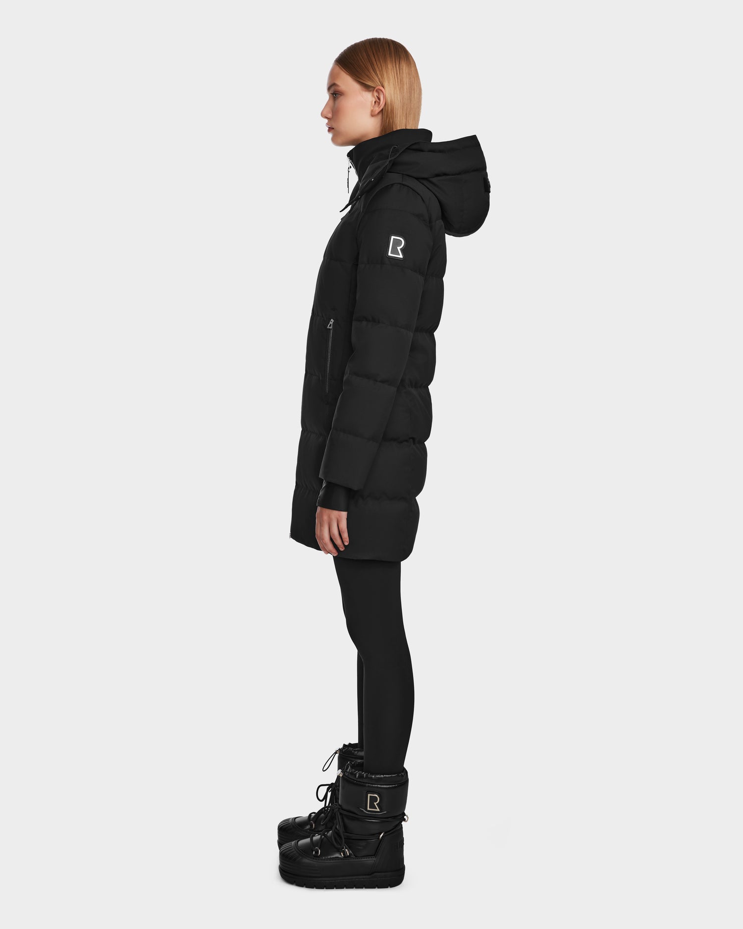 A woman in profile wears Rudsak’s MIRIAM - L123544 Black weather resistant jacket and black winter boots, both with a white R logo, paired with black leggings. The background is plain white.