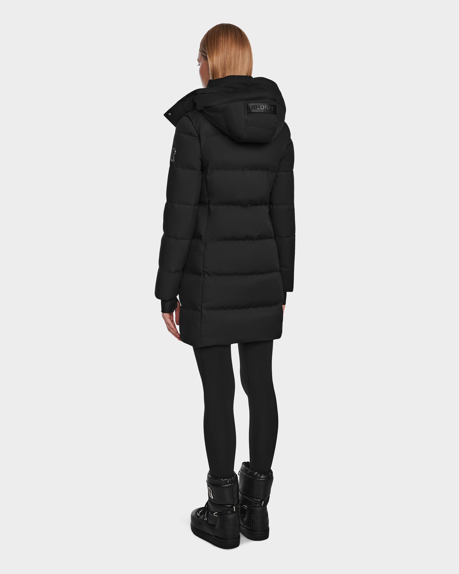 A person with straight, light brown hair wears the Rudsak MIRIAM - L123544 Black womens long down puffer coat with a hood, paired with black leggings and boots, standing against a plain light background, facing away from the camera.