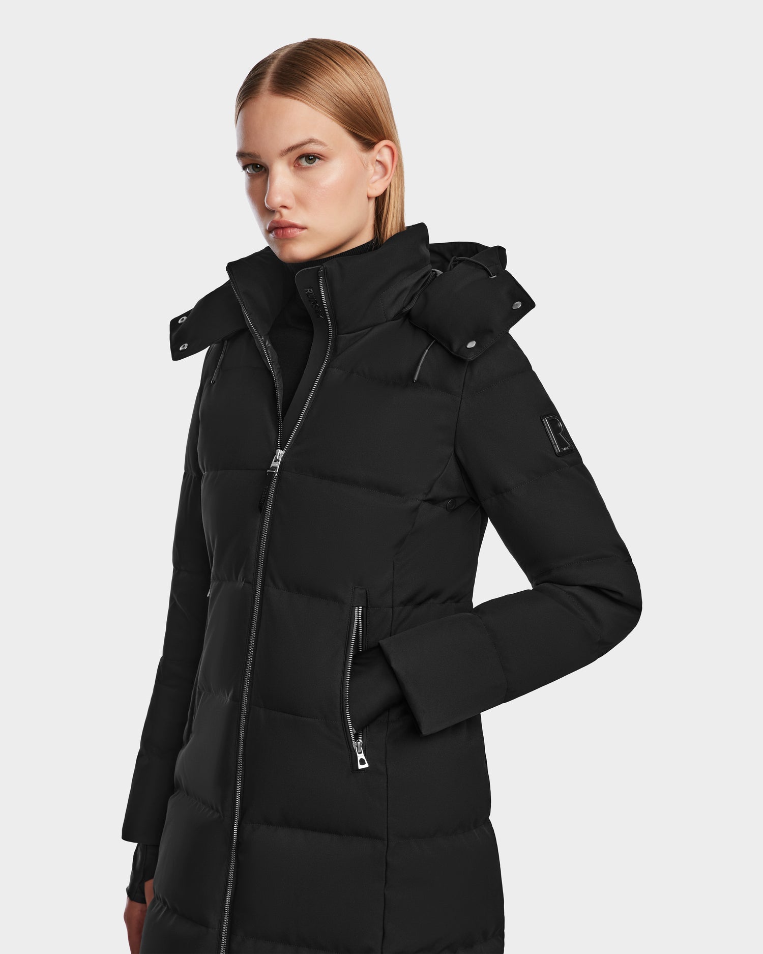 A woman with straight blonde hair models the Rudsak MIRIAM - L123544 Black long womens down puffer coat with a high collar and zip-up front, standing against a plain light gray background.