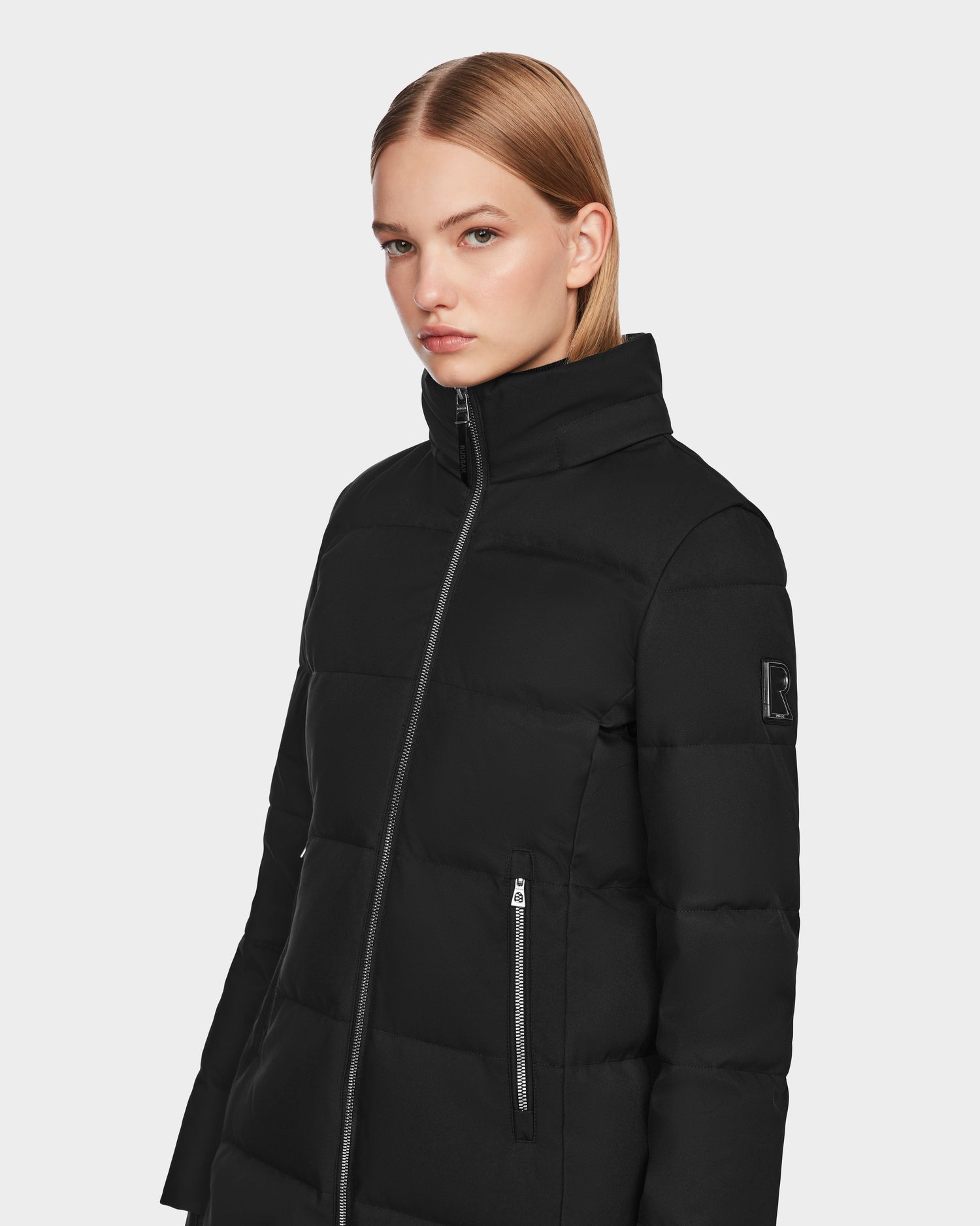 A woman with straight, shoulder-length blonde hair models the Rudsak MIRIAM - L123544 Black women’s down puffer jacket, featuring a high collar and front zipper, against a plain light background.