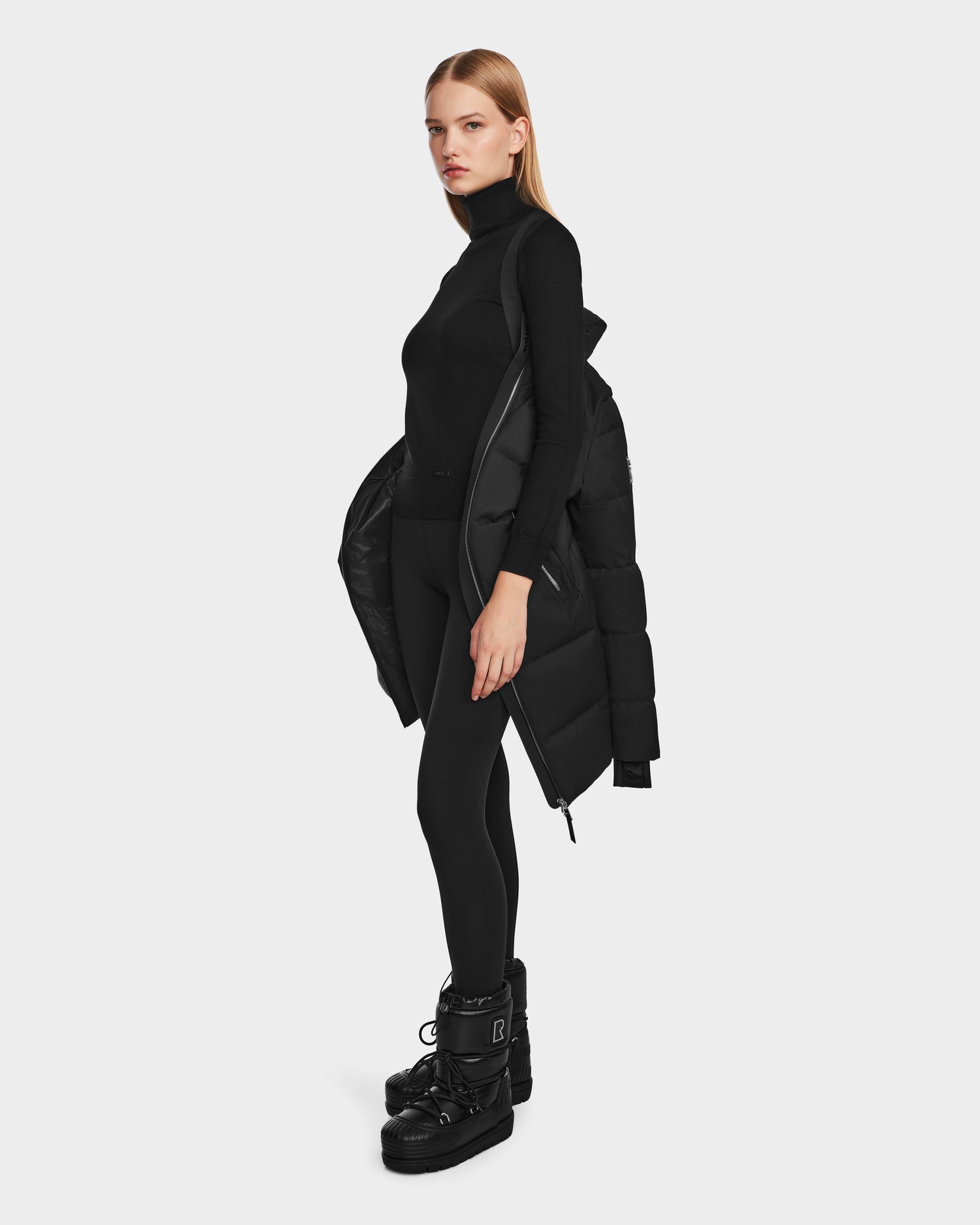 A woman stands in the Rudsak MIRIAM - L123544 Black weather-resistant jacket draped over one shoulder, paired with a black turtleneck, leggings, and chunky boots for a bold winter look against a plain white background.