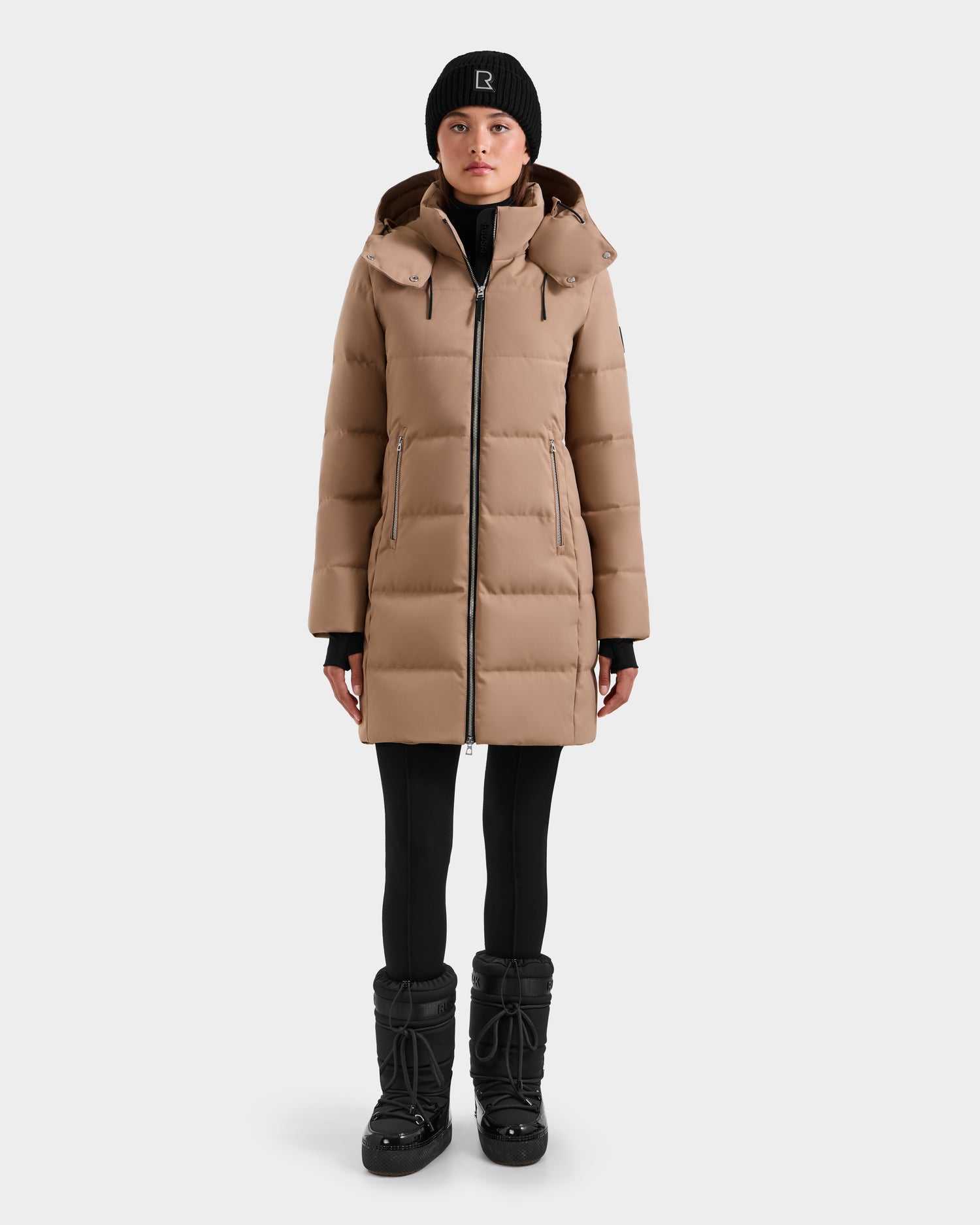 A person models the Rudsak MIRIAM - L123544 Sand women’s down puffer, featuring RDS-certified down, with a beige coat, black beanie, leggings, and winter boots against a plain white background.