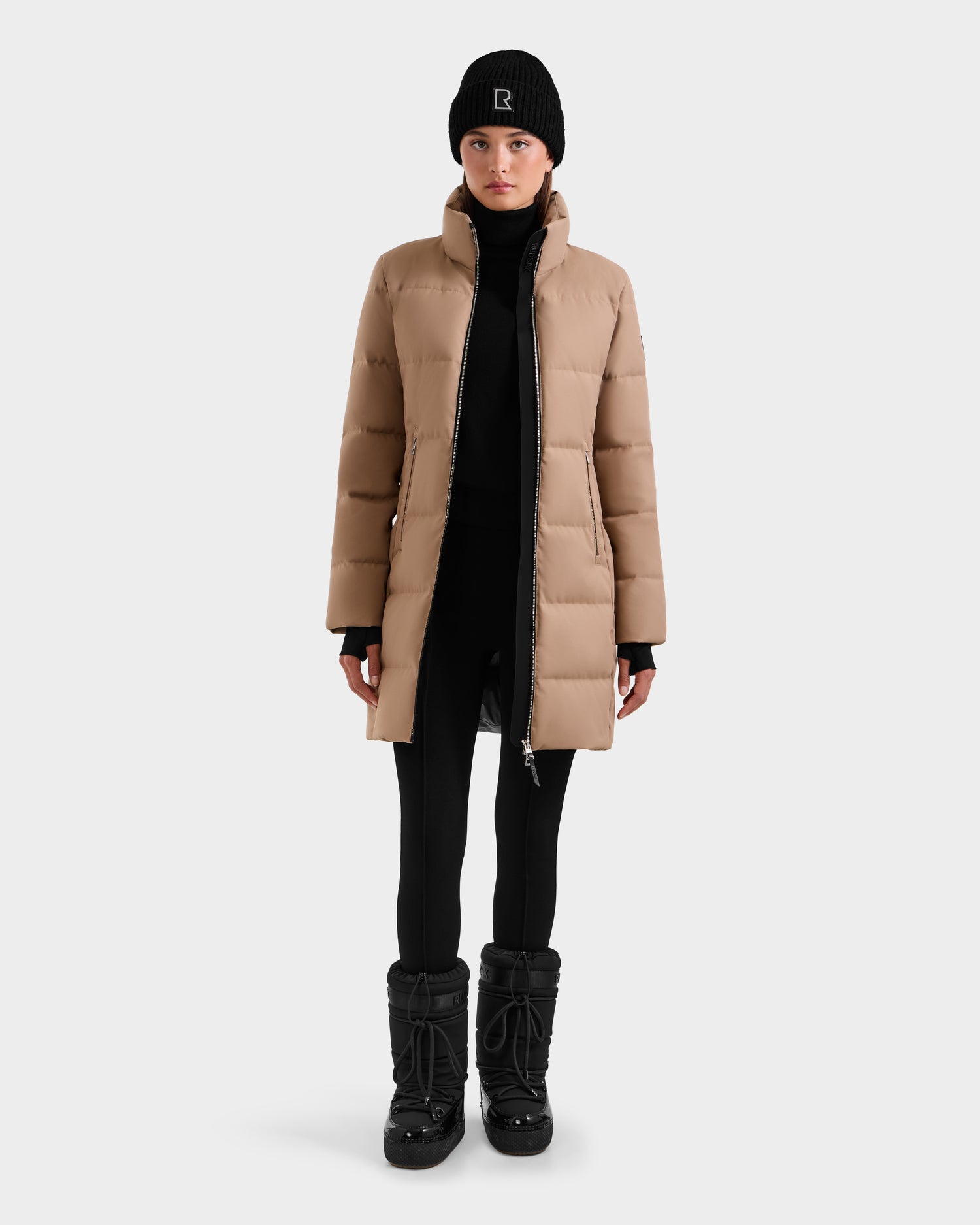 A person wearing the Rudsak MIRIAM - L123544 Sand women's down puffer coat, paired with a black beanie, leggings, and winter boots, stands facing forward against a plain white background.