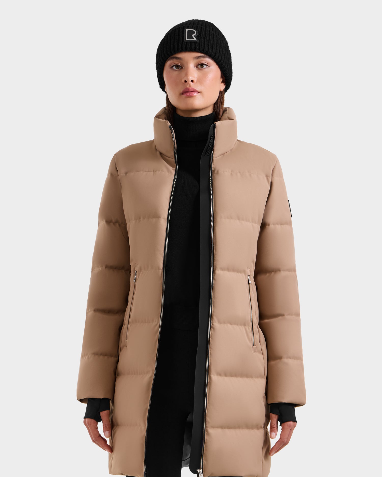 A woman wears Rudsak’s MIRIAM - L123544 Sand, a long beige down puffer coat with zip pockets and high collar, paired with a black turtleneck and R beanie—an RDS-certified winter essential against a plain light background.