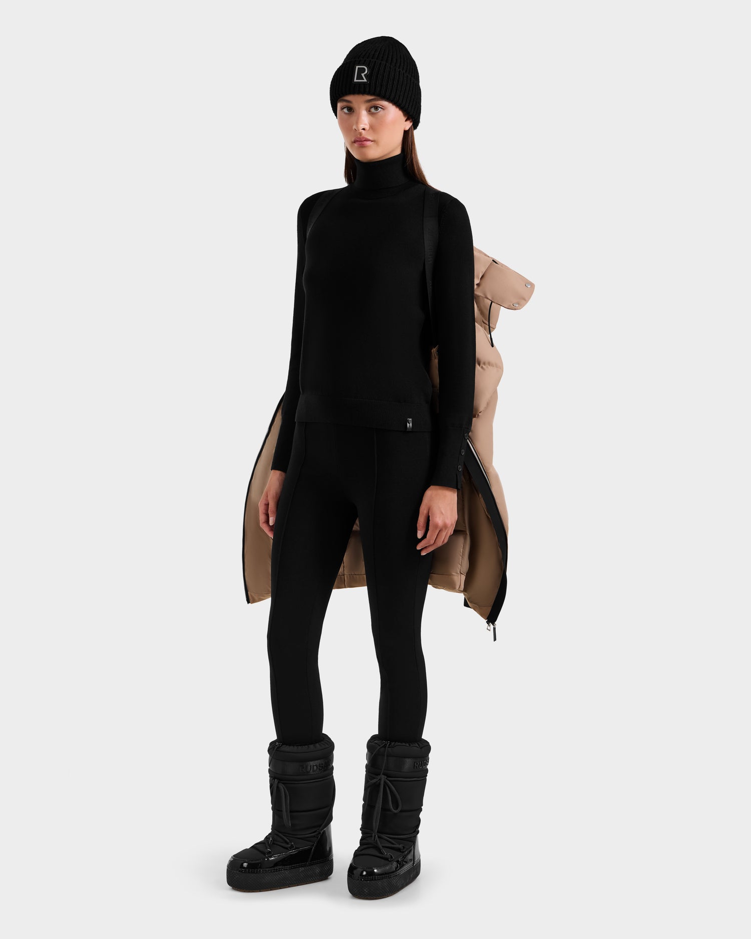 A woman wears a black turtleneck, leggings, winter boots, and a beanie. Draped over her shoulders is the Rudsak MIRIAM - L123544 Sand beige women's down puffer with visible lining. The background is plain white.