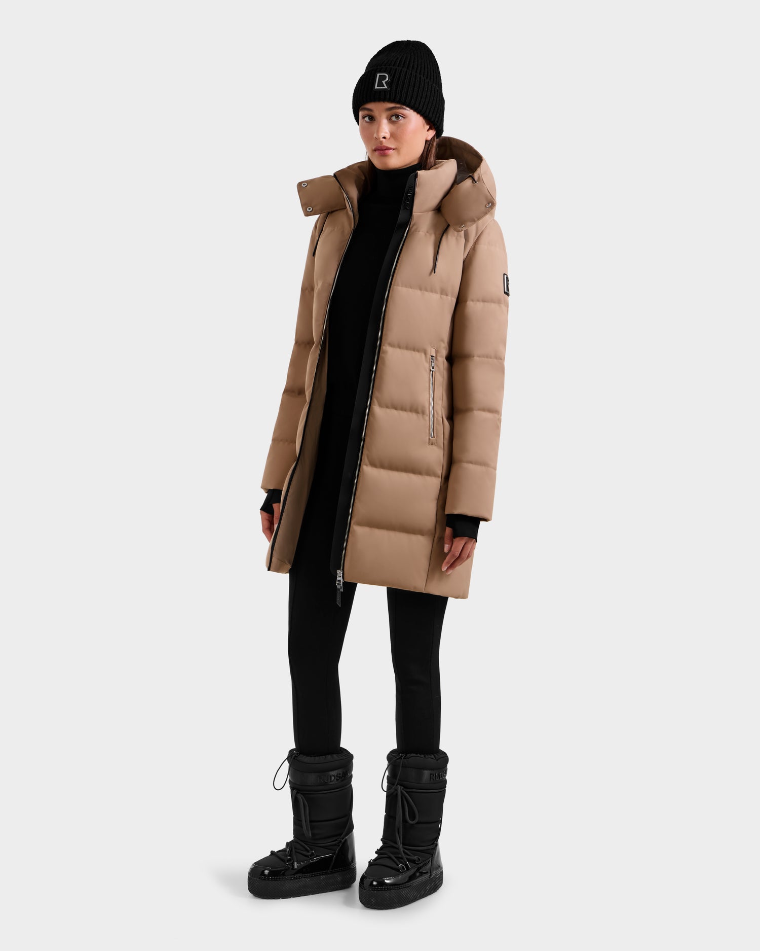 A person wearing the Rudsak MIRIAM - L123544 Sand women's down puffer jacket, black beanie, black leggings, and black winter boots stands against a plain light grey background.