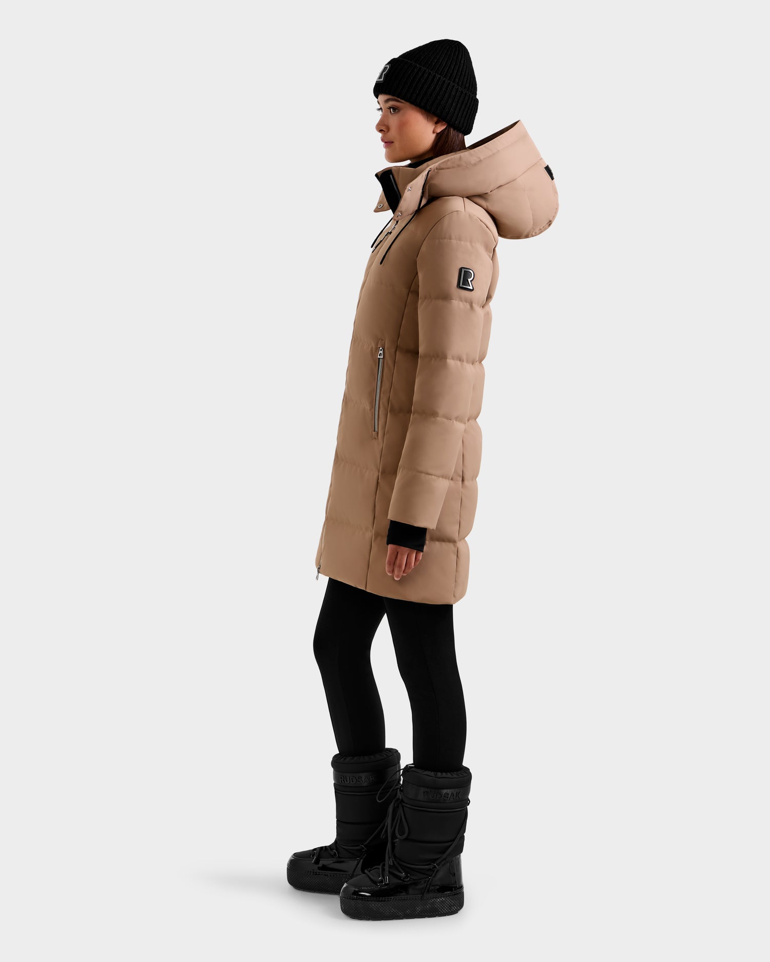 A person wearing the Rudsak MIRIAM - L123544 Sand women's down puffer with a hood, black leggings, winter boots, and a black knit beanie stands facing left against a plain light background.
