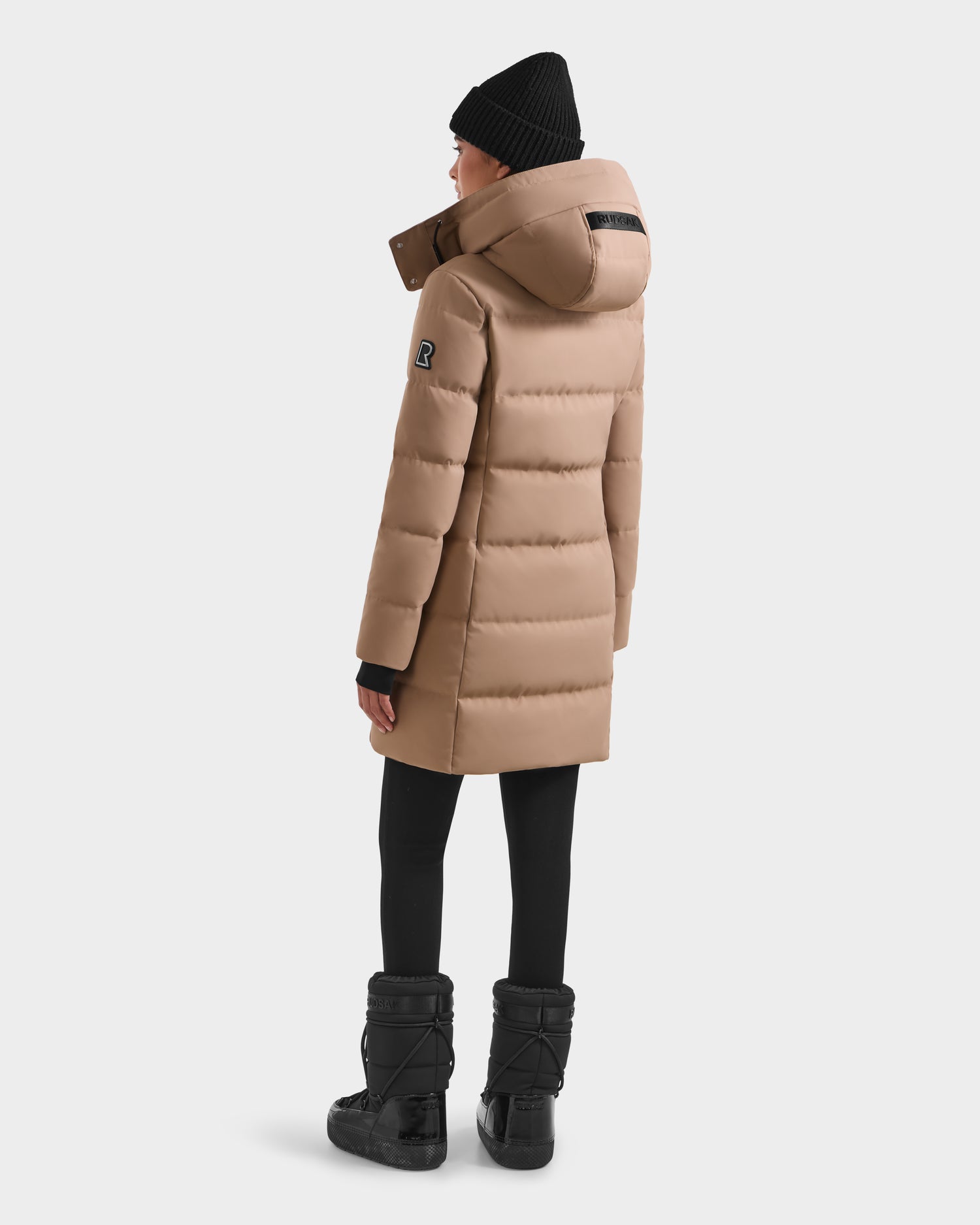 A person stands facing away against a plain white background, wearing the Rudsak MIRIAM - L123544 Sand women's down puffer coat with a hood, paired with a black beanie, black leggings, and winter boots.