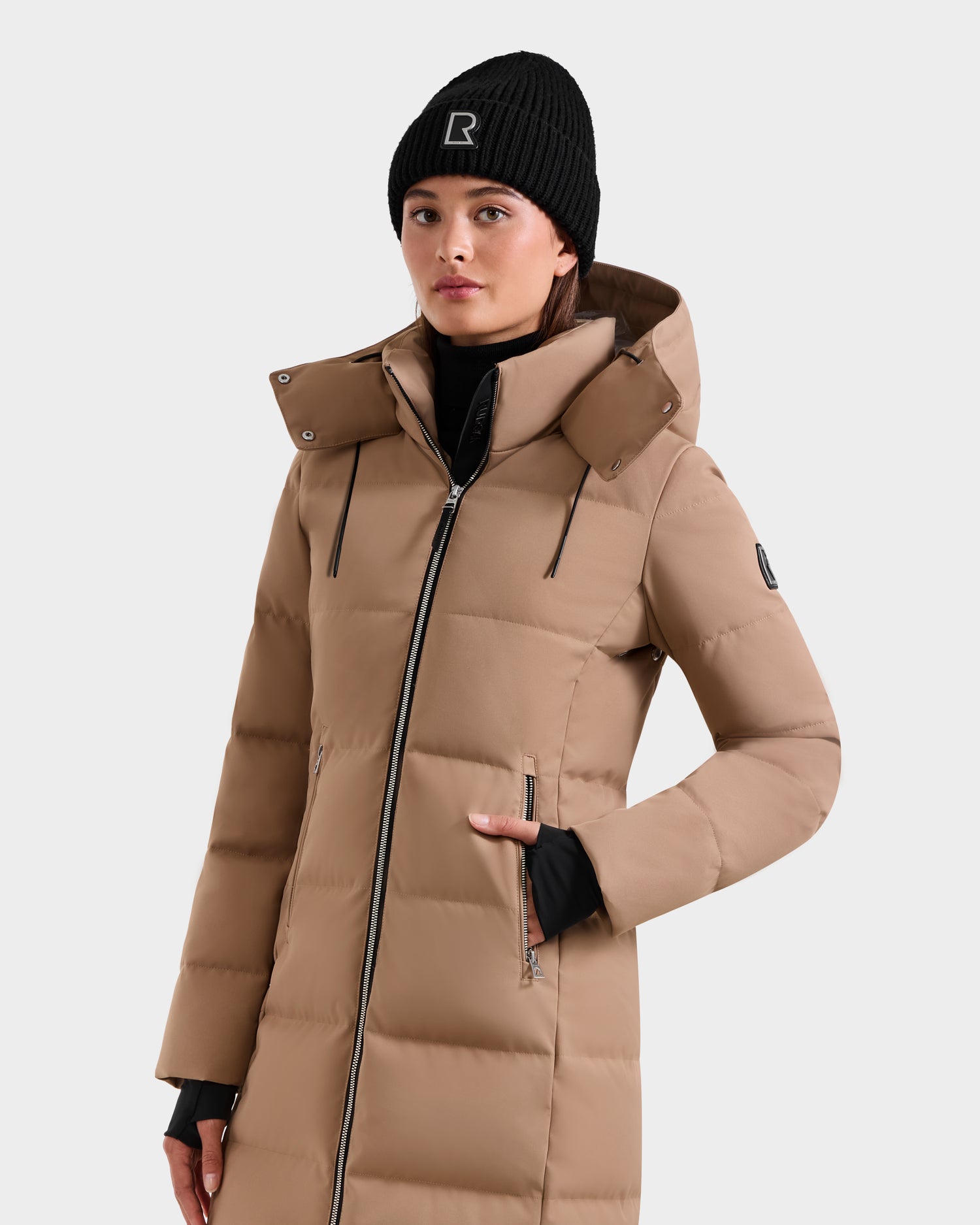 A person wears the Rudsak MIRIAM - L123544 Sand women’s down puffer coat with black zippers and RDS-certified down, paired with a black knit beanie featuring a white logo, against a plain light background.