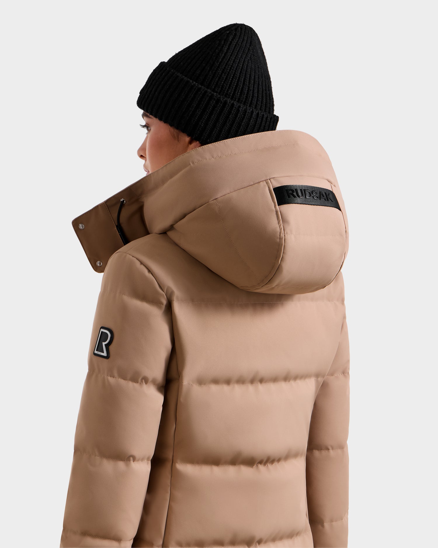 A person wears the Rudsak MIRIAM - L123544 Sand women's down puffer jacket with a large hood and black beanie, featuring a black Rudsak patch on the hood, shown from the back and side while facing away from the camera.