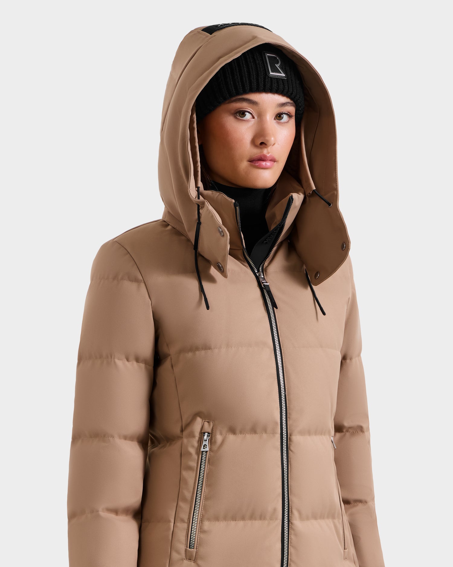 A person wearing the Rudsak MIRIAM - L123544 Sand women's down puffer jacket with a hood and a black beanie, standing against a plain light background and facing forward with a neutral expression.