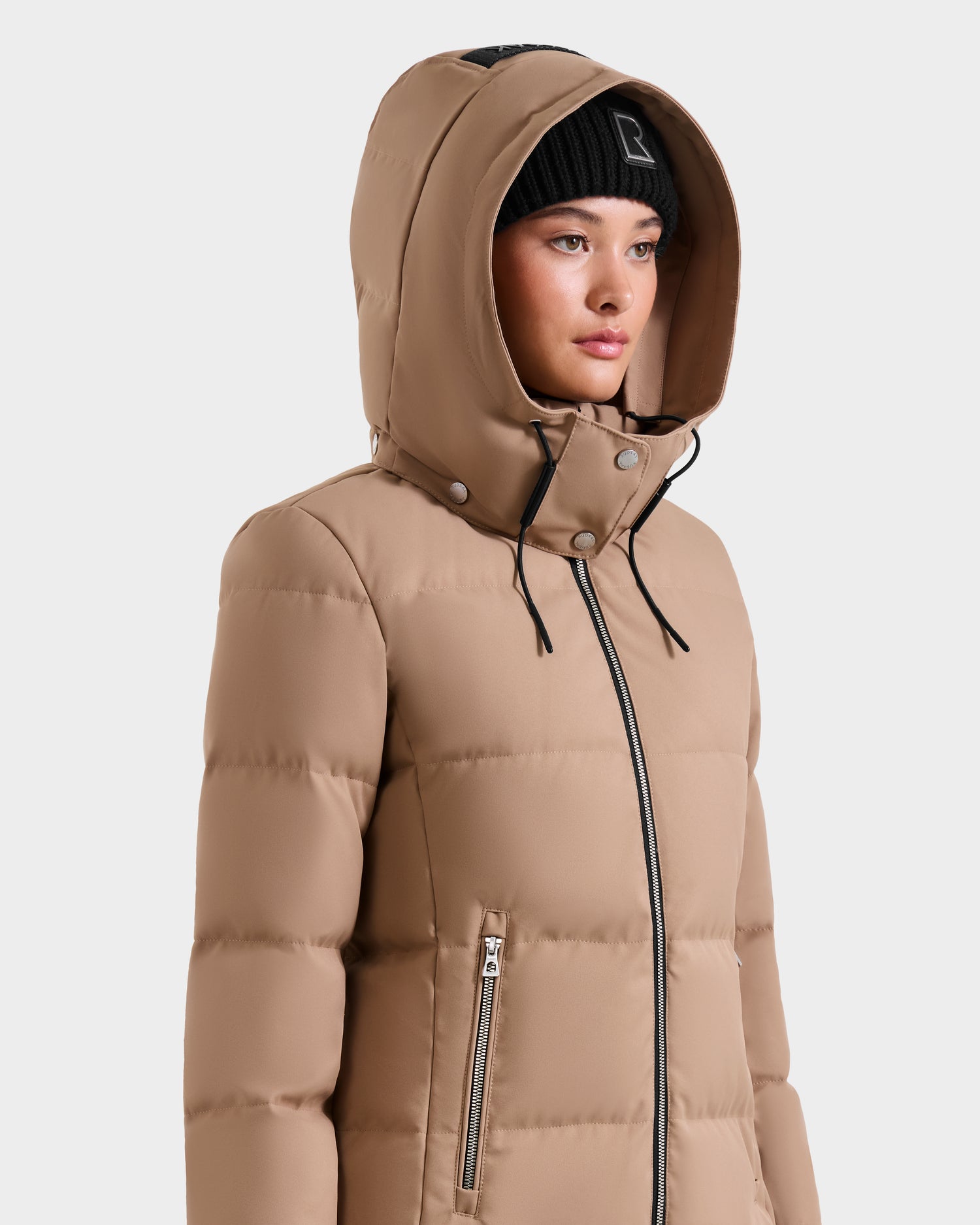 A young woman wears the Rudsak MIRIAM - L123544 Sand RDS-certified down puffer jacket with hood and a black beanie, standing against a plain white background and looking slightly to the side.