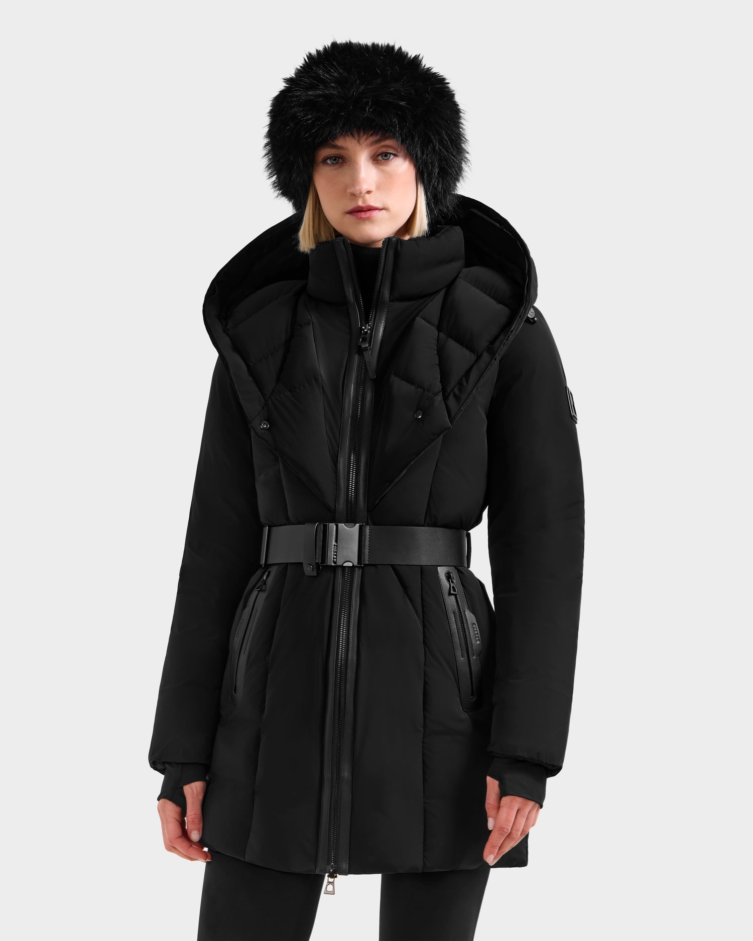 A woman wears the Rudsak MODA FF - 8125969 Black/Black belted winter coat with a large faux fur hood and matching faux fur hat, standing against a plain light background.
