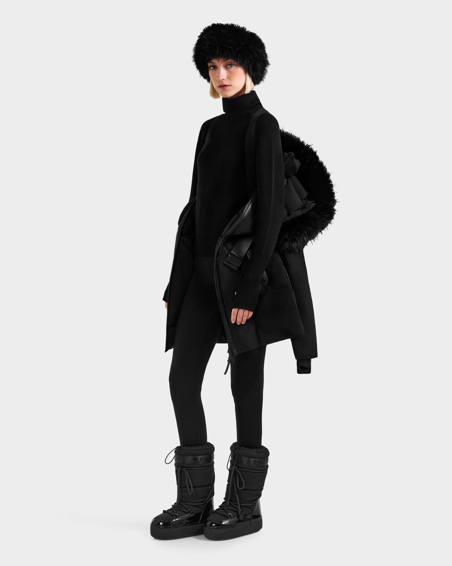 A person in an all-black winter look wears the Rudsak MODA FF - 8125969 Black/Black fitted down puffer with a chic faux fur hood, paired with a turtleneck, fitted pants, fur hat, and chunky snow boots against a plain light background.